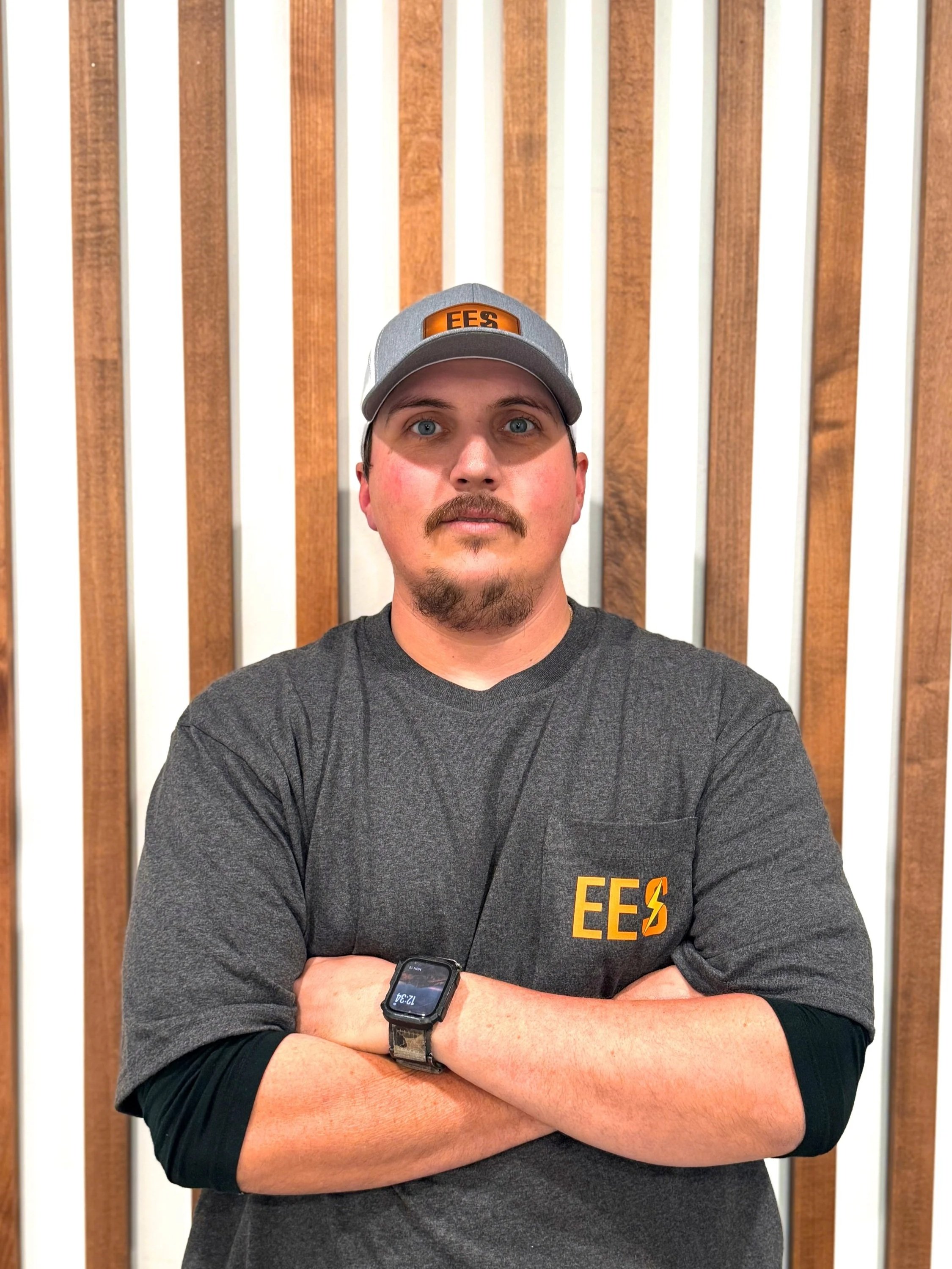 A man with crossed arms wearing a gray cap and a dark gray shirt with orange and yellow "EES" logo, standing in front of a wooden wall.