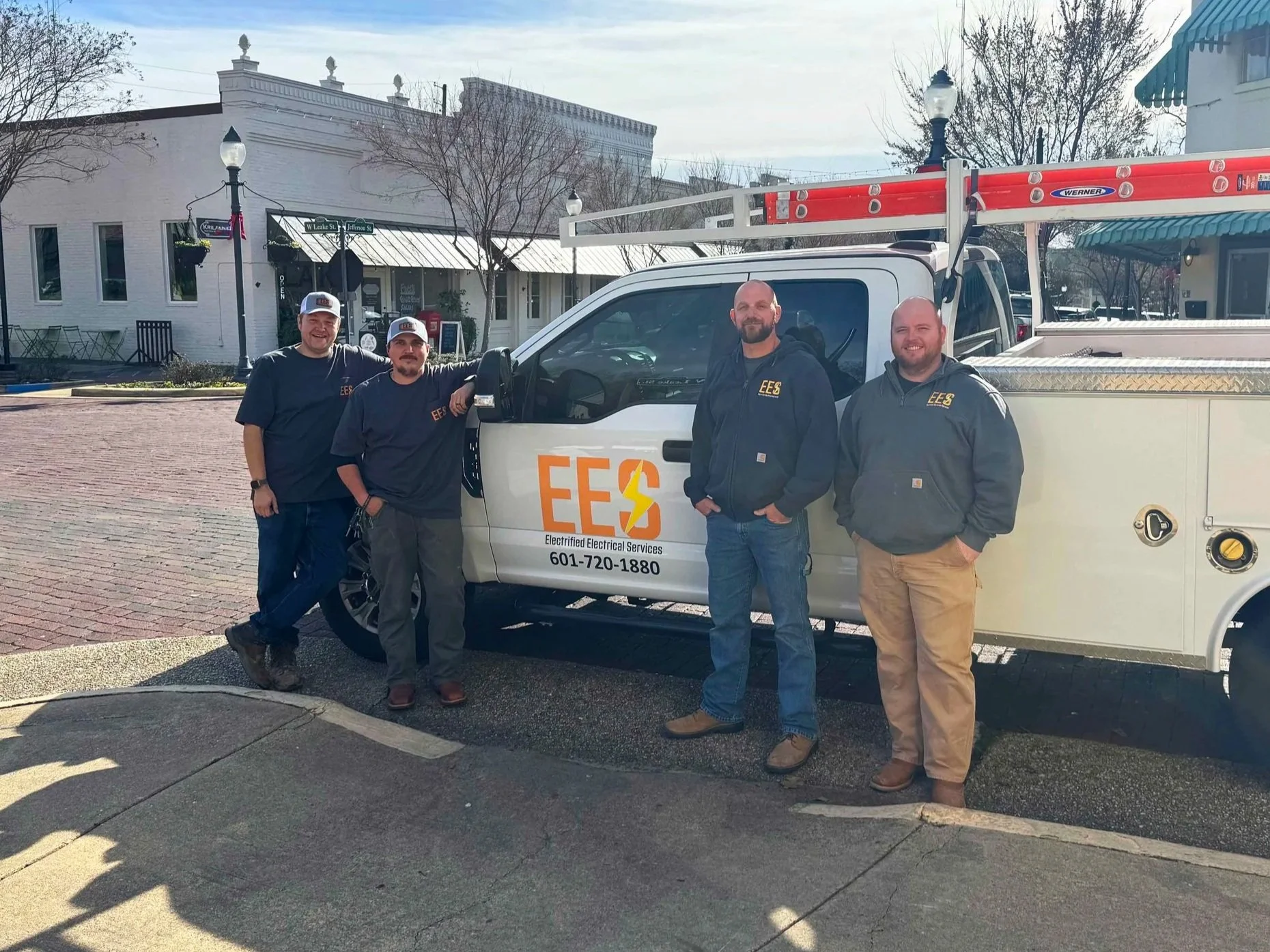 Four men standing in front of a white vehicle with the logo and contact info of Electrified Electrical Services, with buildings and trees in the background.