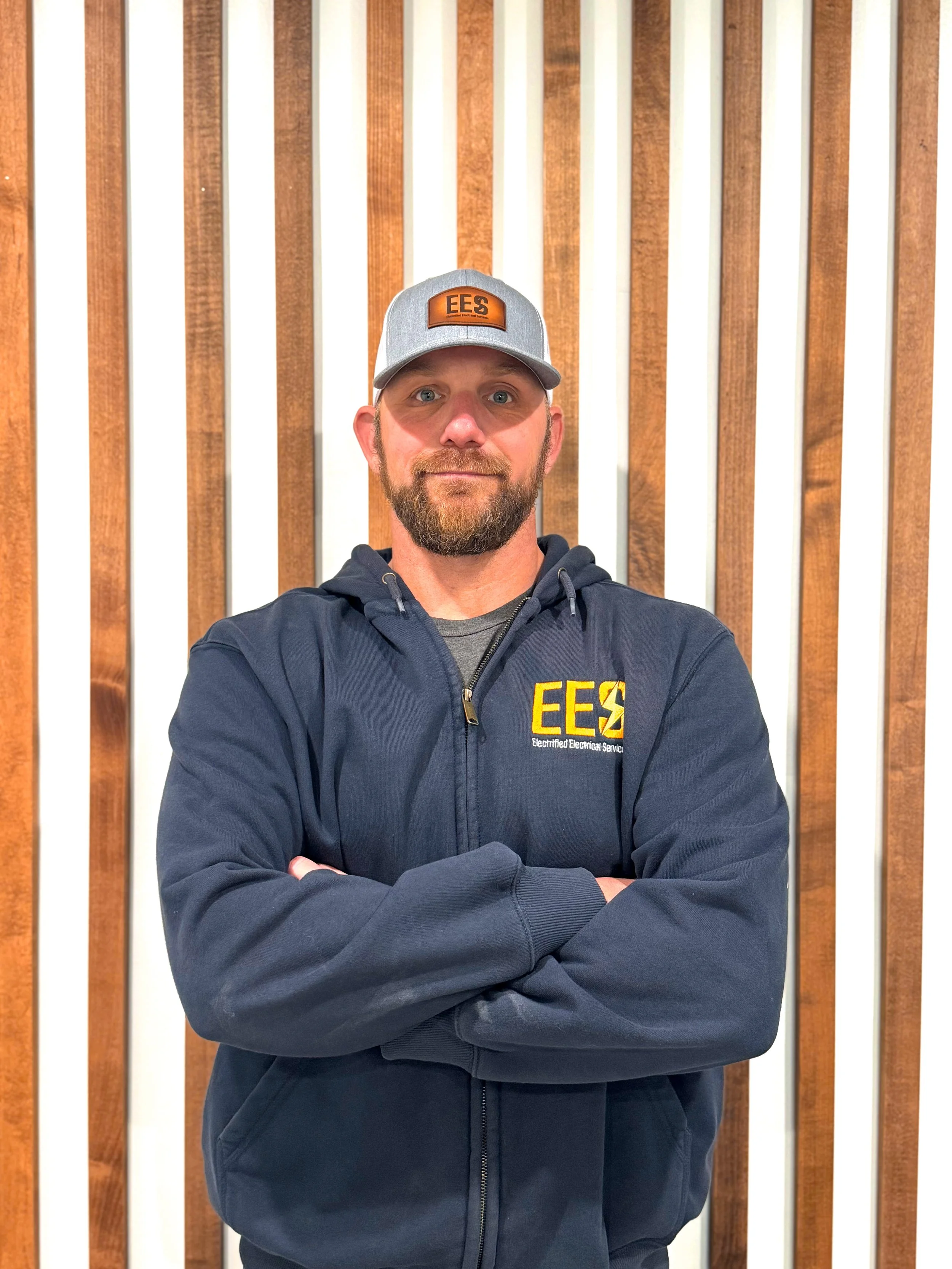 A man with a beard and blue eyes wearing a gray cap with an orange patch labeled 'EES' and a navy blue hoodie with yellow 'EES' logo, crossed arms, standing in front of a wooden wall.