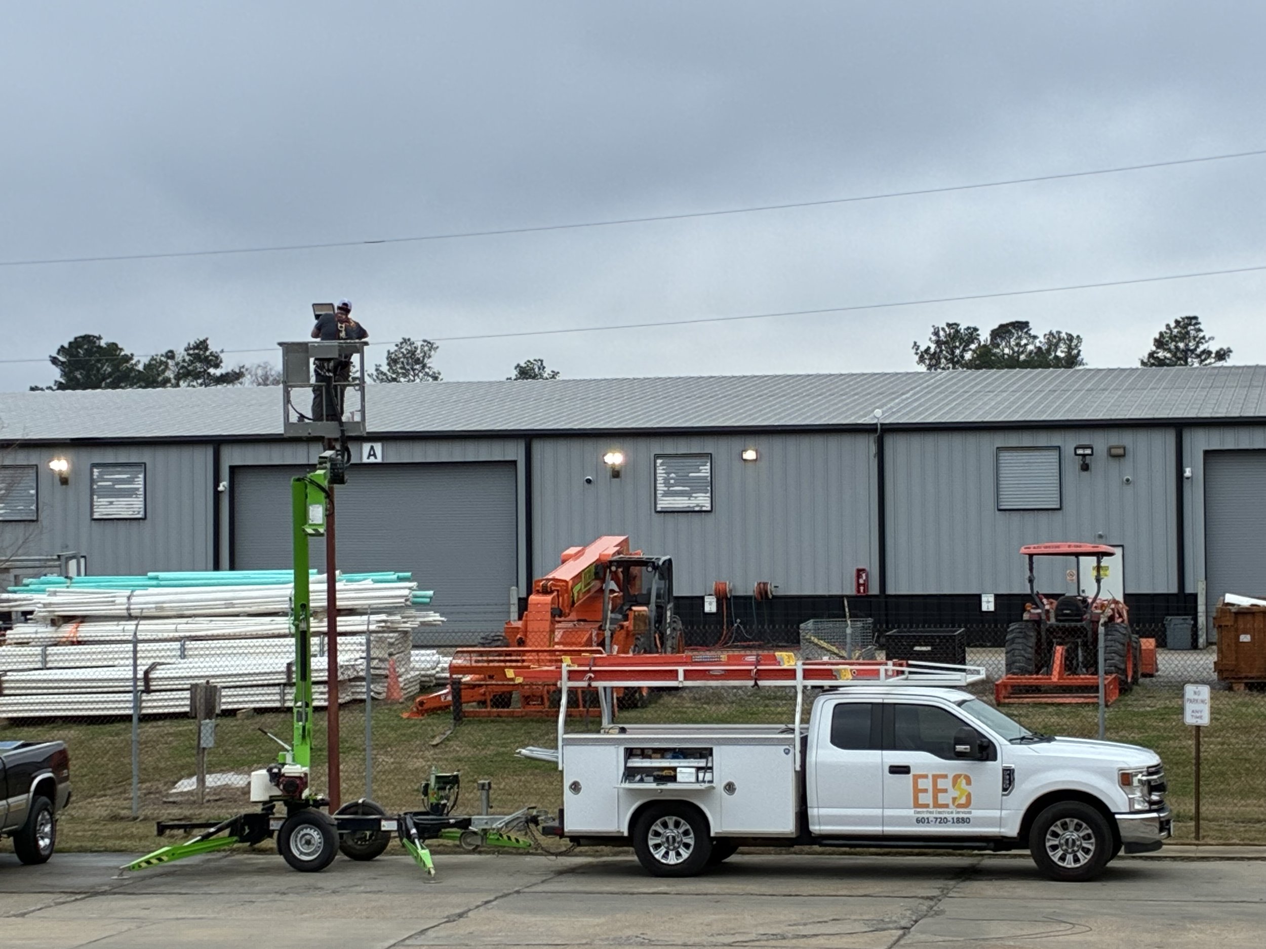A maintenance worker working on a streetlight from a raised lift platform, with utility vehicles and construction equipment parked outside a warehouse.