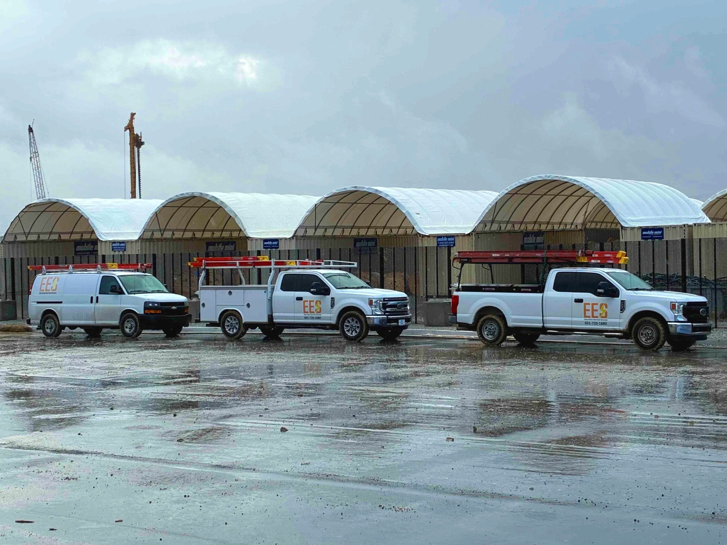 Three utility trucks parked in front of a covered walkway under gray, overcast sky and wet pavement.