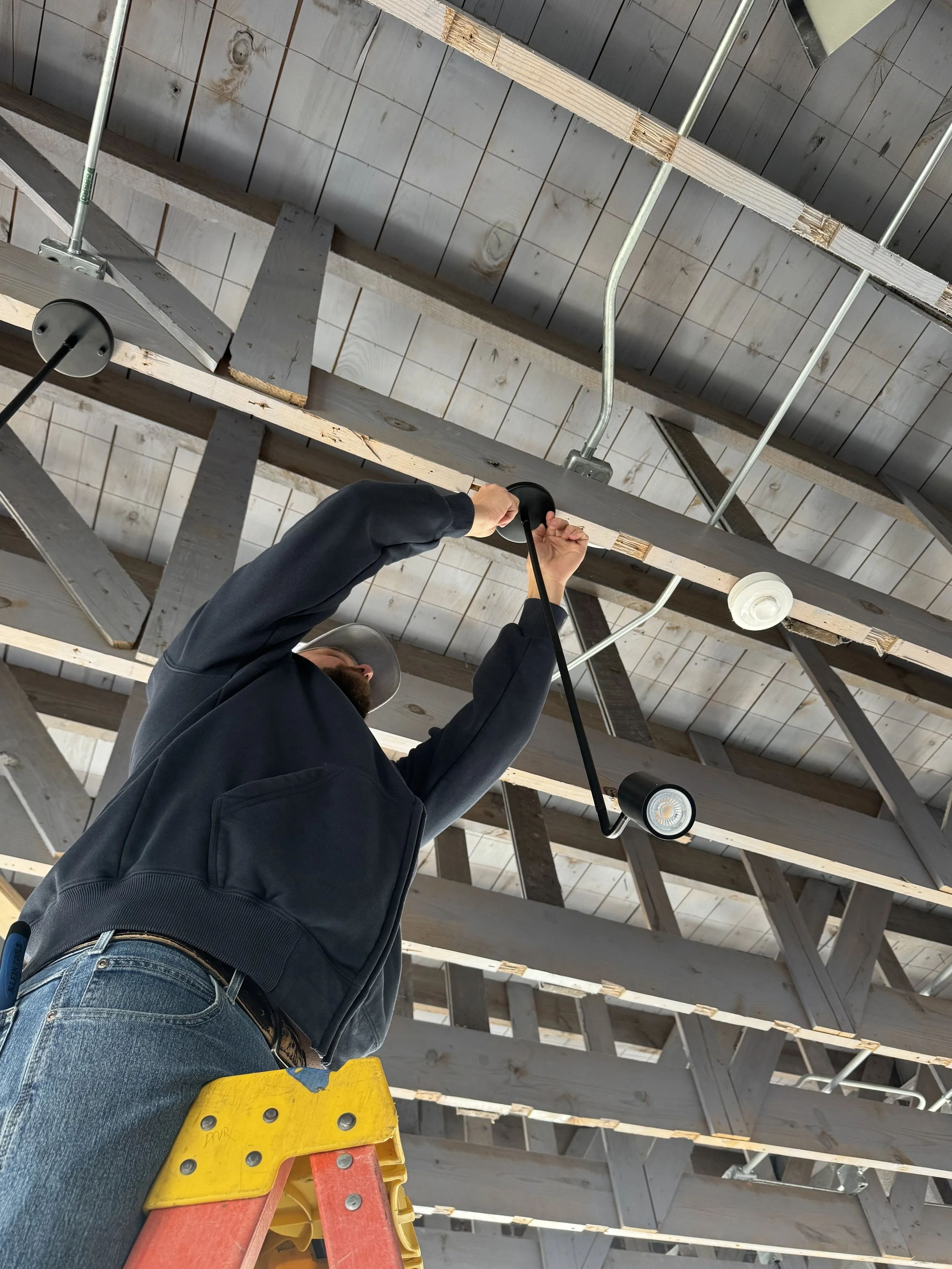A man standing on a ladder installing or adjusting a light fixture on a wooden ceiling in a building under construction.