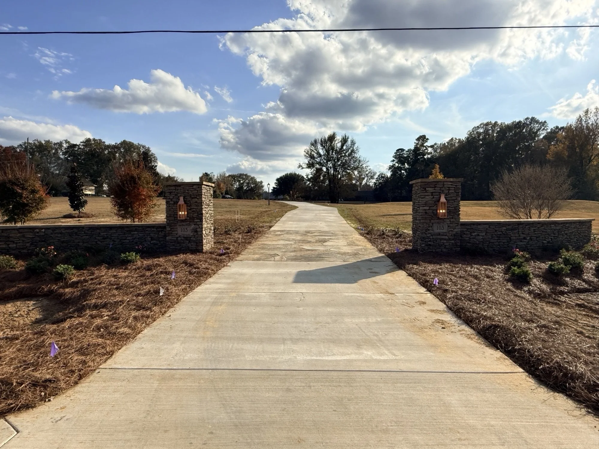 A view of a concrete driveway entrance with two stone pillars on each side, each with a lantern. The driveway extends into a grassy area with trees and bushes, under a partly cloudy sky.