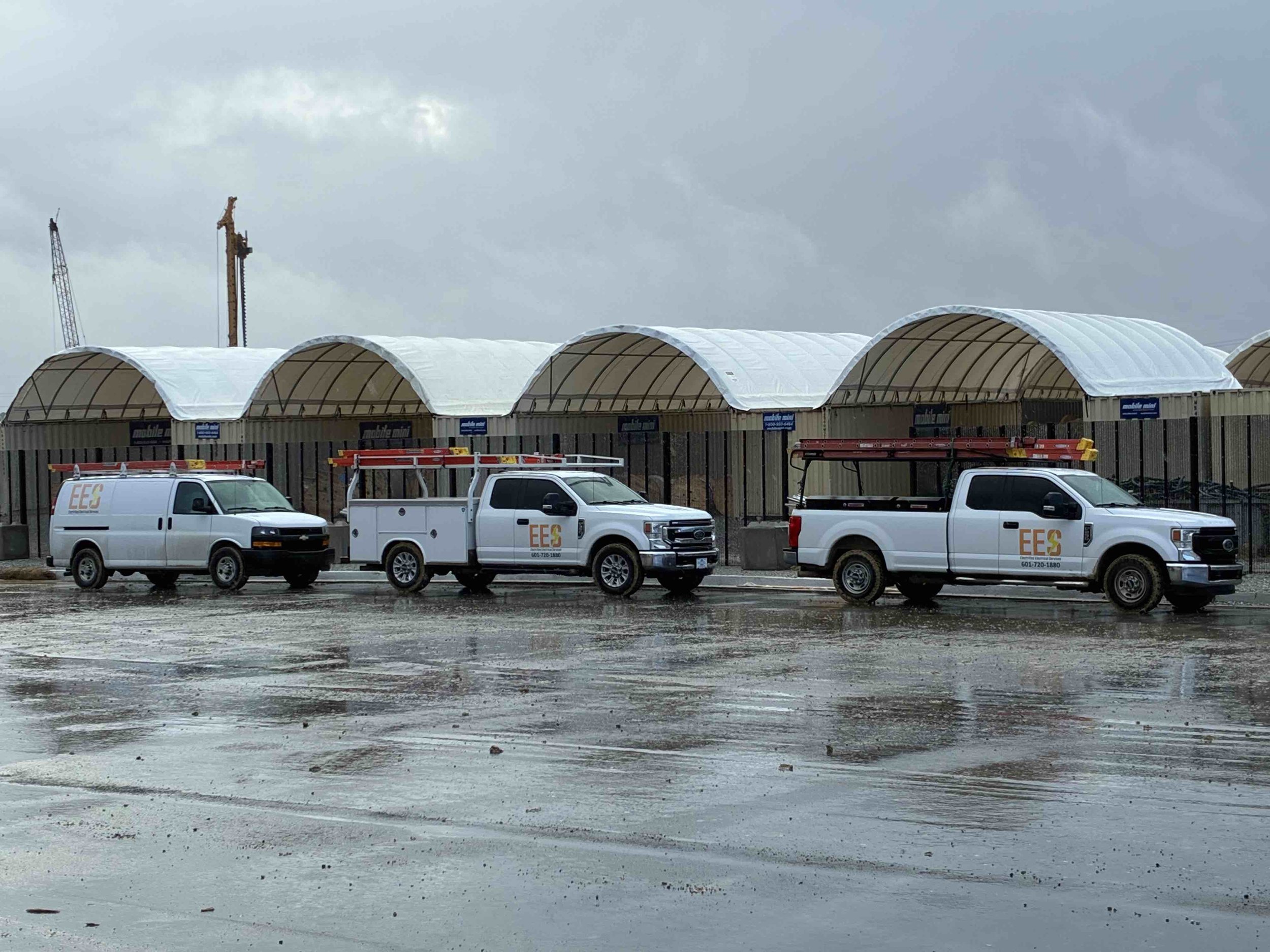 Three utility trucks parked in front of a commercial building with a curved roof structure. The trucks are white with orange and gray branding, and each is carrying ladders or equipment. The ground is wet, indicating recent rain, and the sky is cloud
