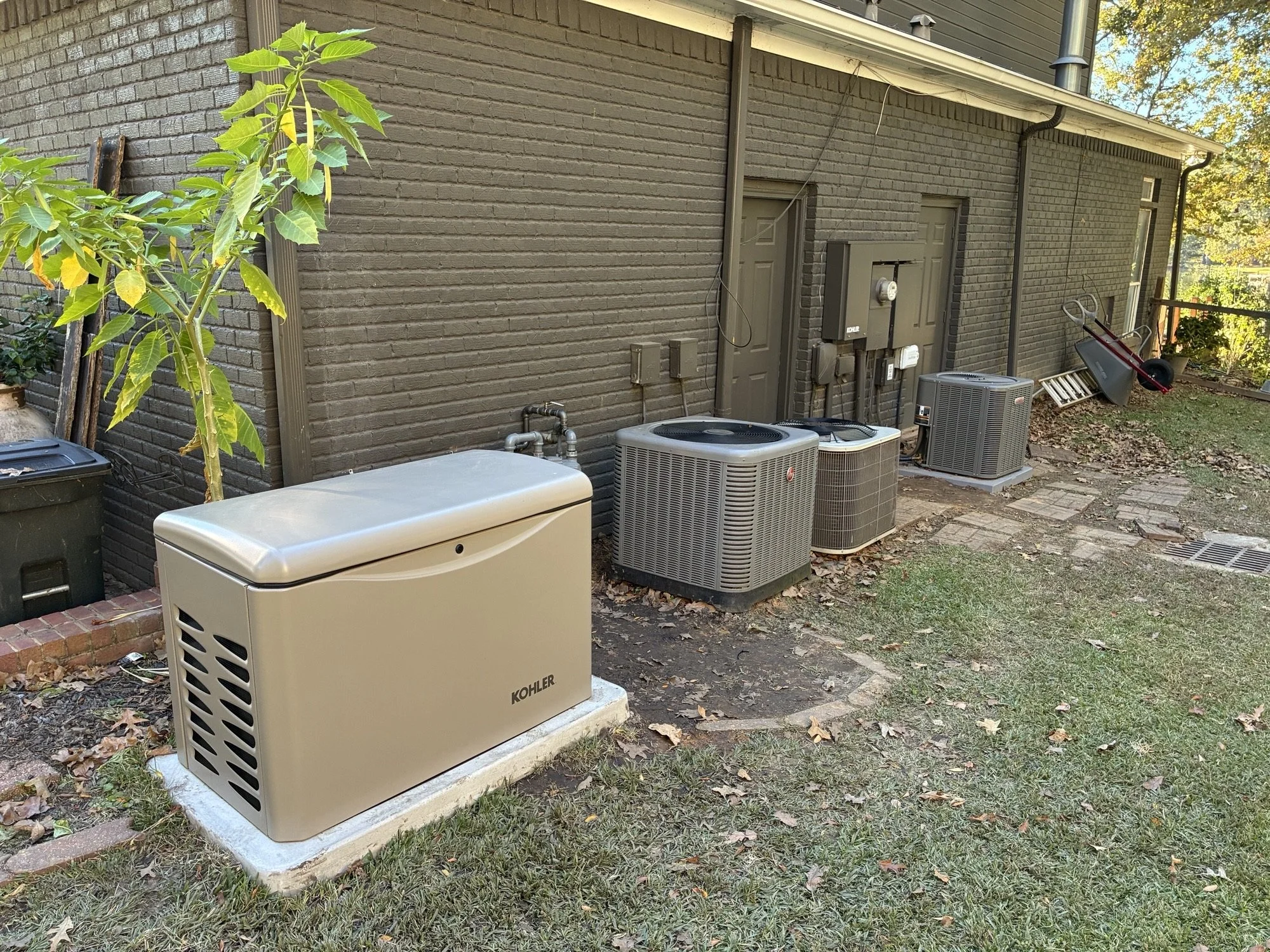 Backyard with multiple air conditioning units and a Kohler generator installed against a brick house wall. There is a wheelbarrow, a ladder, and some leaves on the ground.