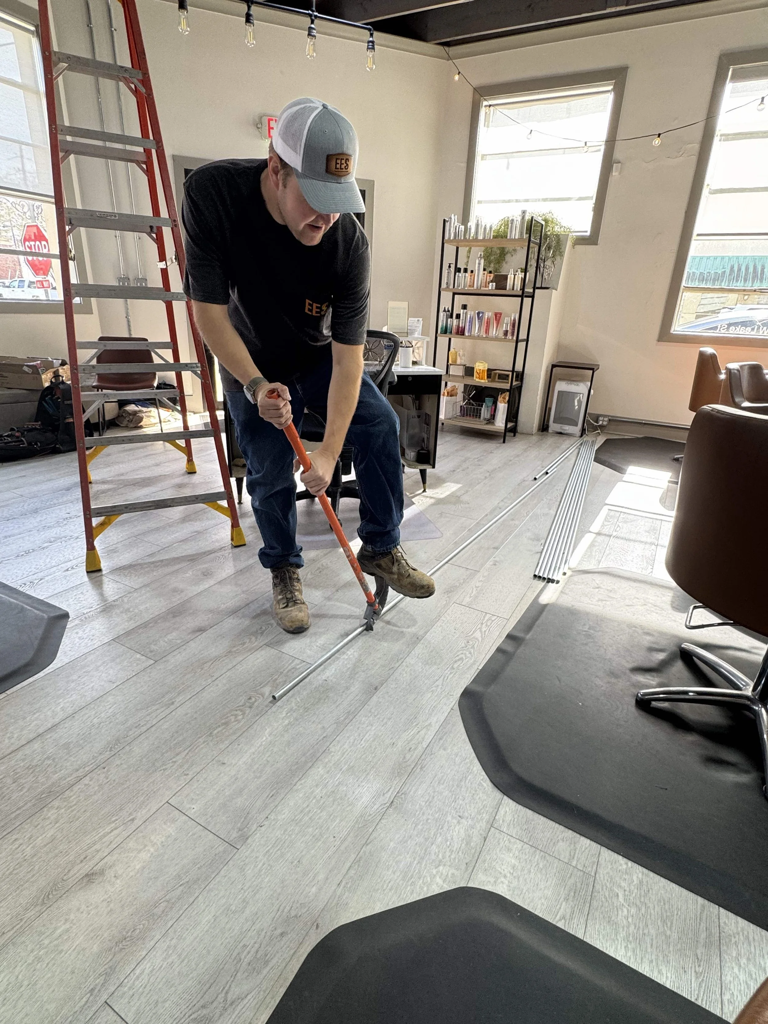 A man wearing a gray trucker hat with 'EES' logo, black shirt, and jeans is using a pipe cutter on a metal pipe on a light-colored wooden floor in a room with large windows, a ladder, and shelves.
