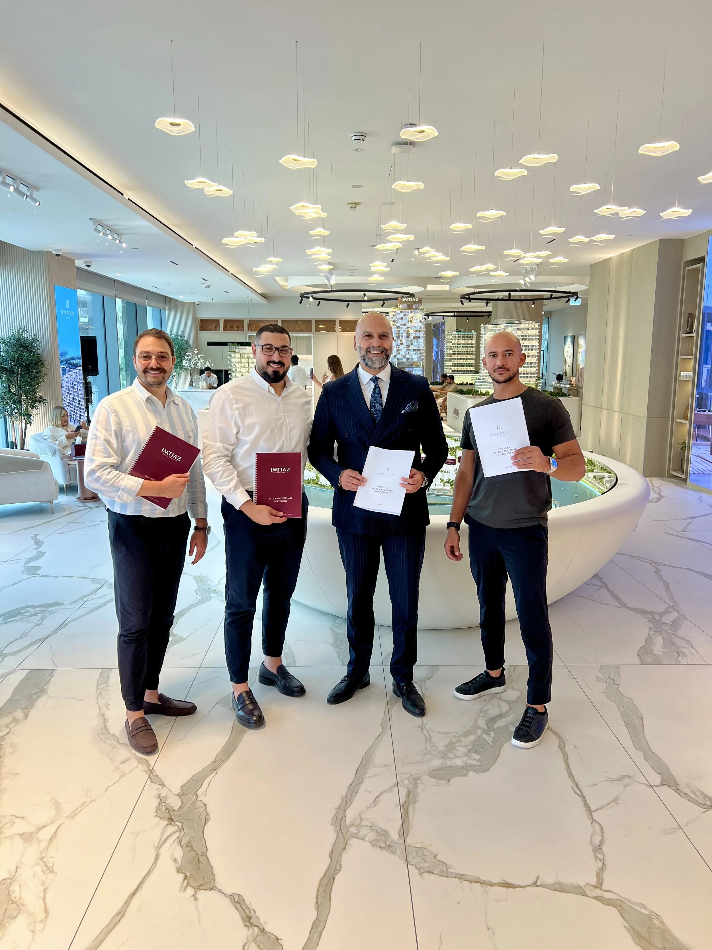 Four men standing together in a modern office lobby, holding certificates. One man in a suit is in the middle, with a beard and smiling, flanked by three men in casual attire. The lobby has marble floors and contemporary light fixtures.