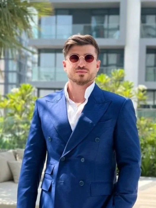 A man in a blue suit and white shirt wearing sunglasses, standing outdoors with modern glass buildings and greenery in the background.