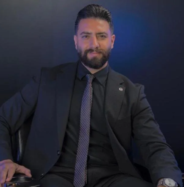 A man with dark hair and a beard wearing a dark suit, black shirt, and patterned tie, seated and smiling in front of a dark background.