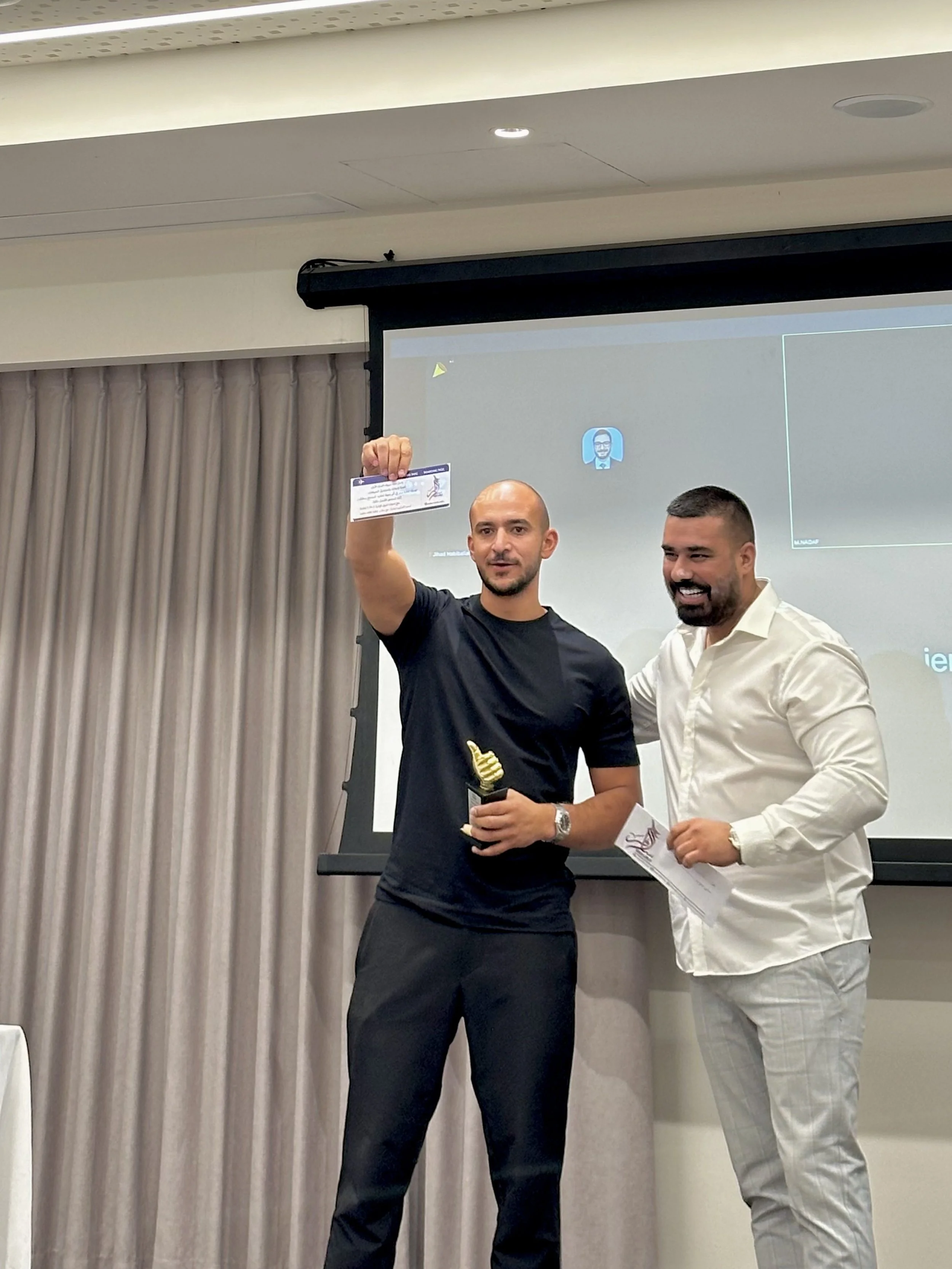 Two men standing in front of a presentation screen, one holding a certificate and an award, smiling during an award ceremony.