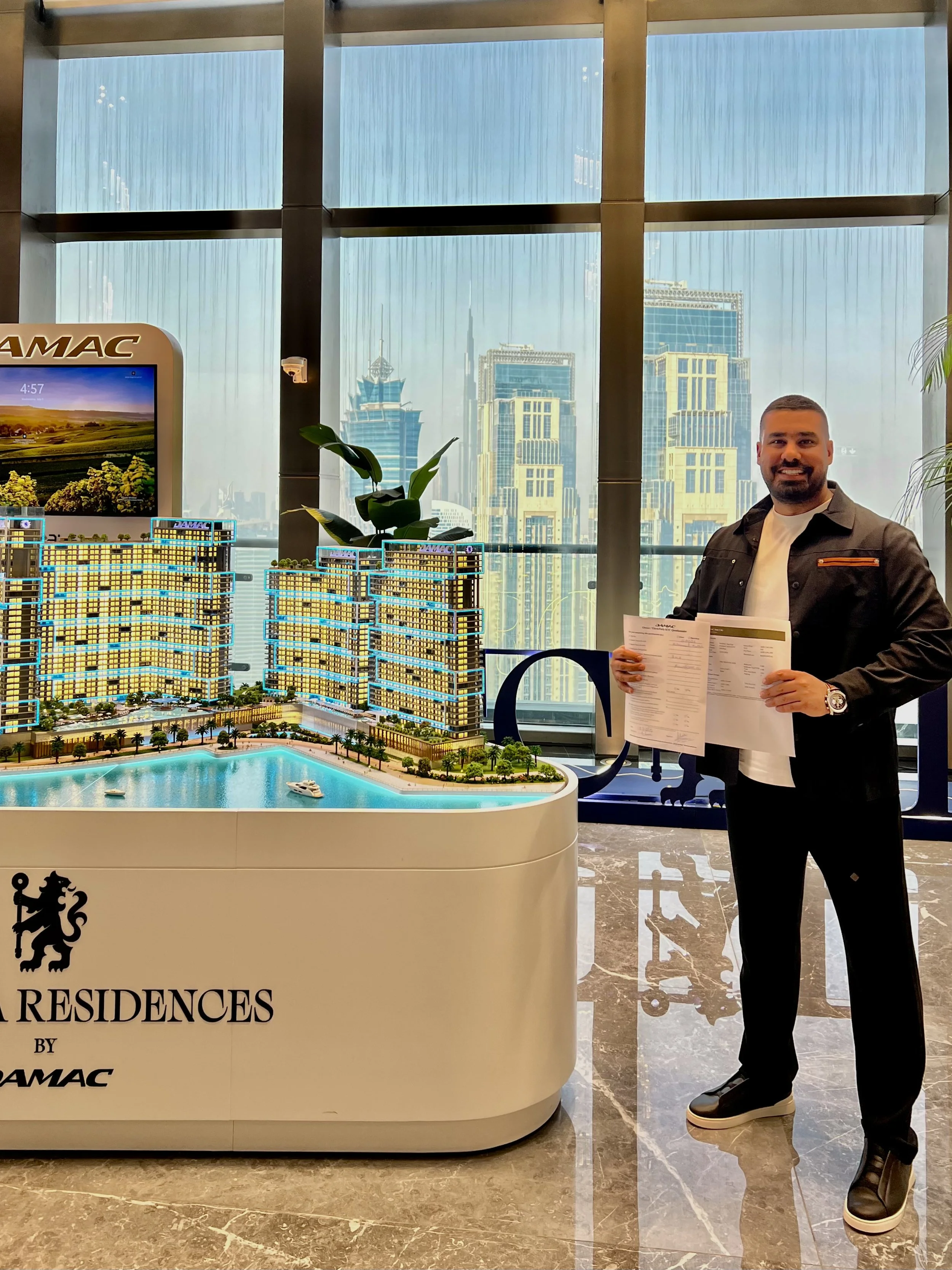 A man holding documents standing next to a scale model of a residential building development, in front of a city skyline with tall buildings, inside a high-rise office with large windows.