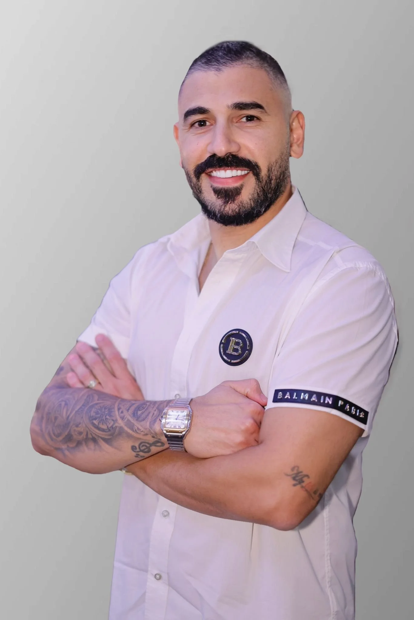 A smiling man with a beard and tattoos on his arms, wearing a white shirt with a Balmain Paris patch on the sleeve, standing with arms crossed against a plain background.