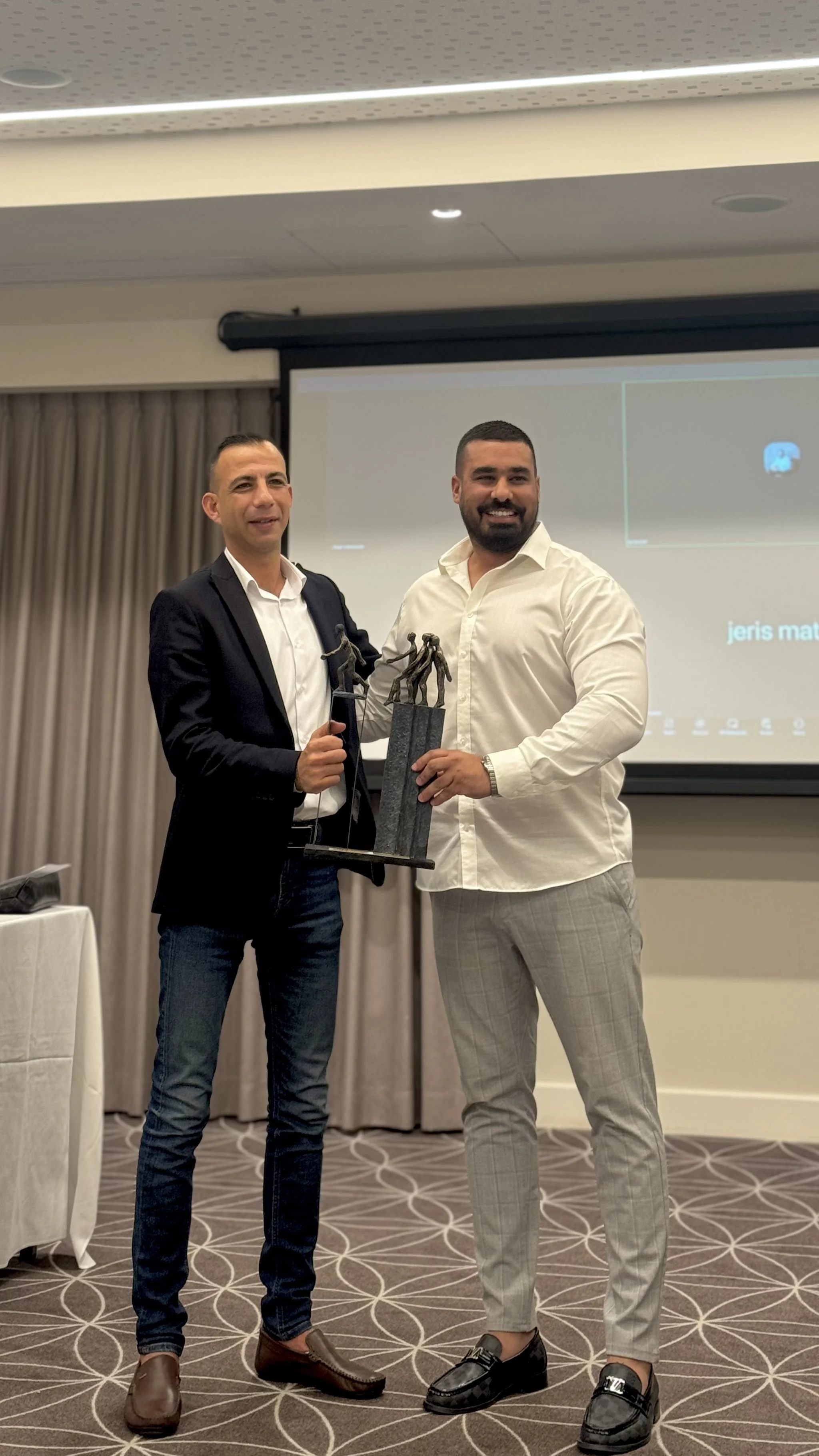 Two men in formal attire standing in front of a projector screen, holding an award or trophy, smiling for the camera, in a conference room or event space.