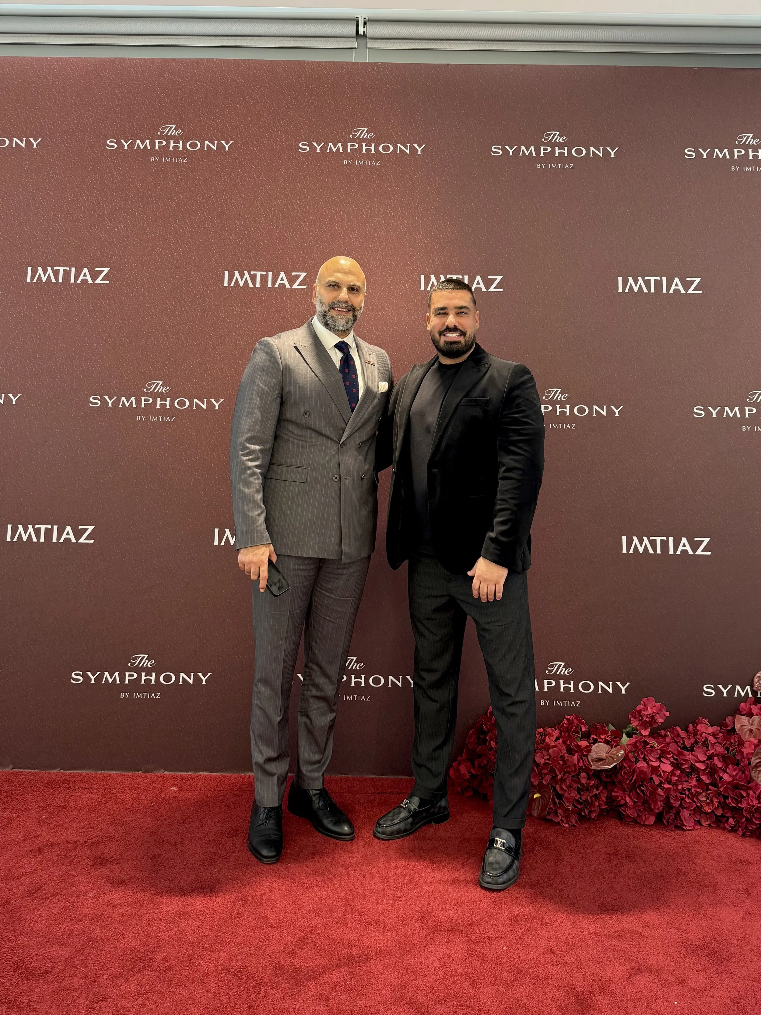 Two men in suits standing in front of a backdrop that reads 'The Symphony by Imtiaz.' One man is wearing a grey pinstripe suit with a tie, and the other man is wearing a black suit. There are red flowers on the floor beside them.