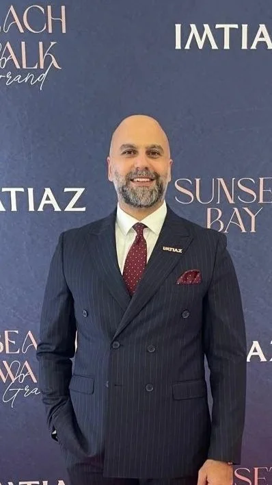 A man in a dark pinstripe suit, white shirt, and red tie, standing in front of a dark blue background with various words and logos, smiling at the camera.
