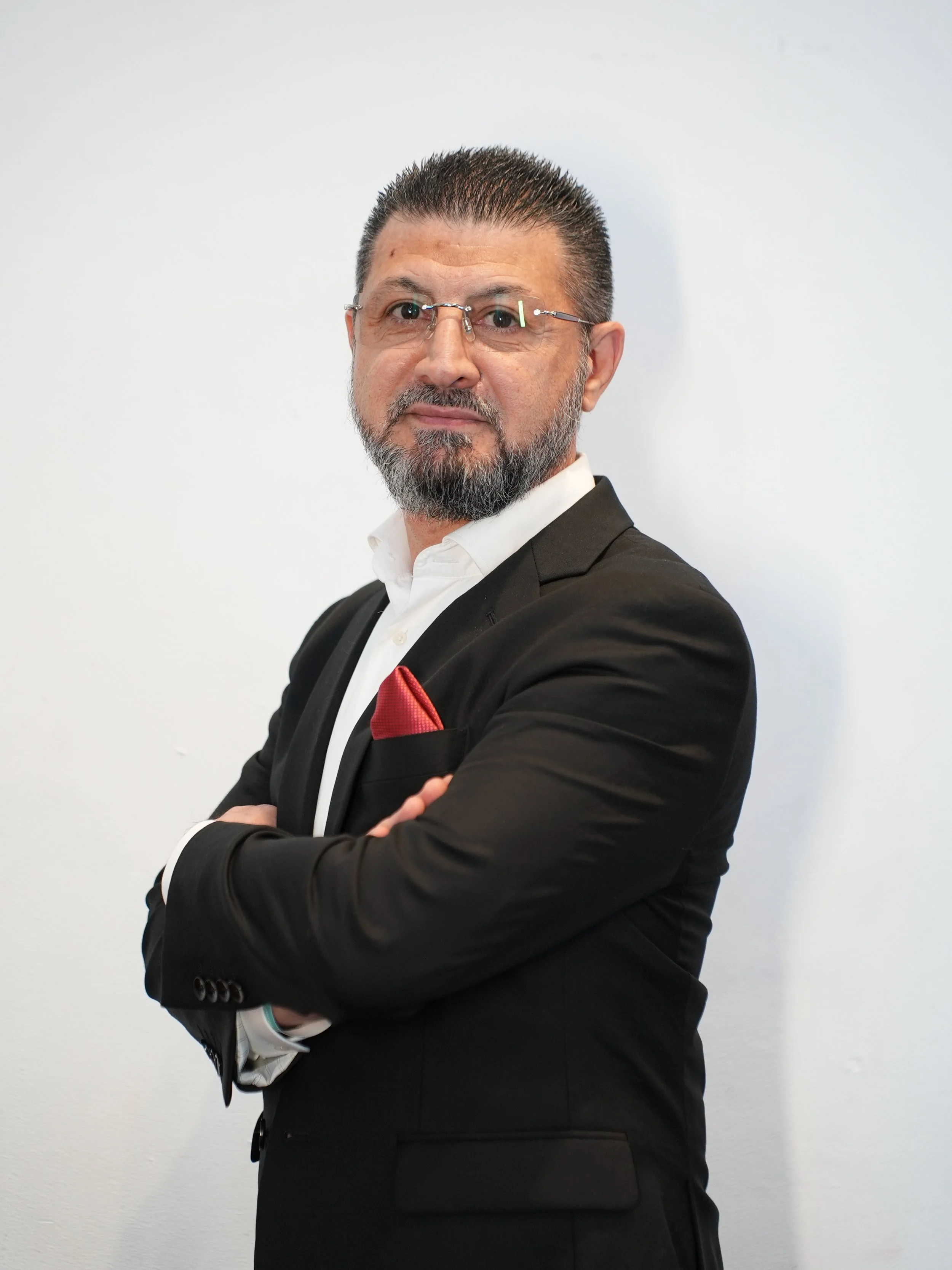 A middle-aged man with glasses, a beard, and short dark hair, wearing a black suit with a white shirt and a red pocket square, standing against a plain white wall with arms crossed.