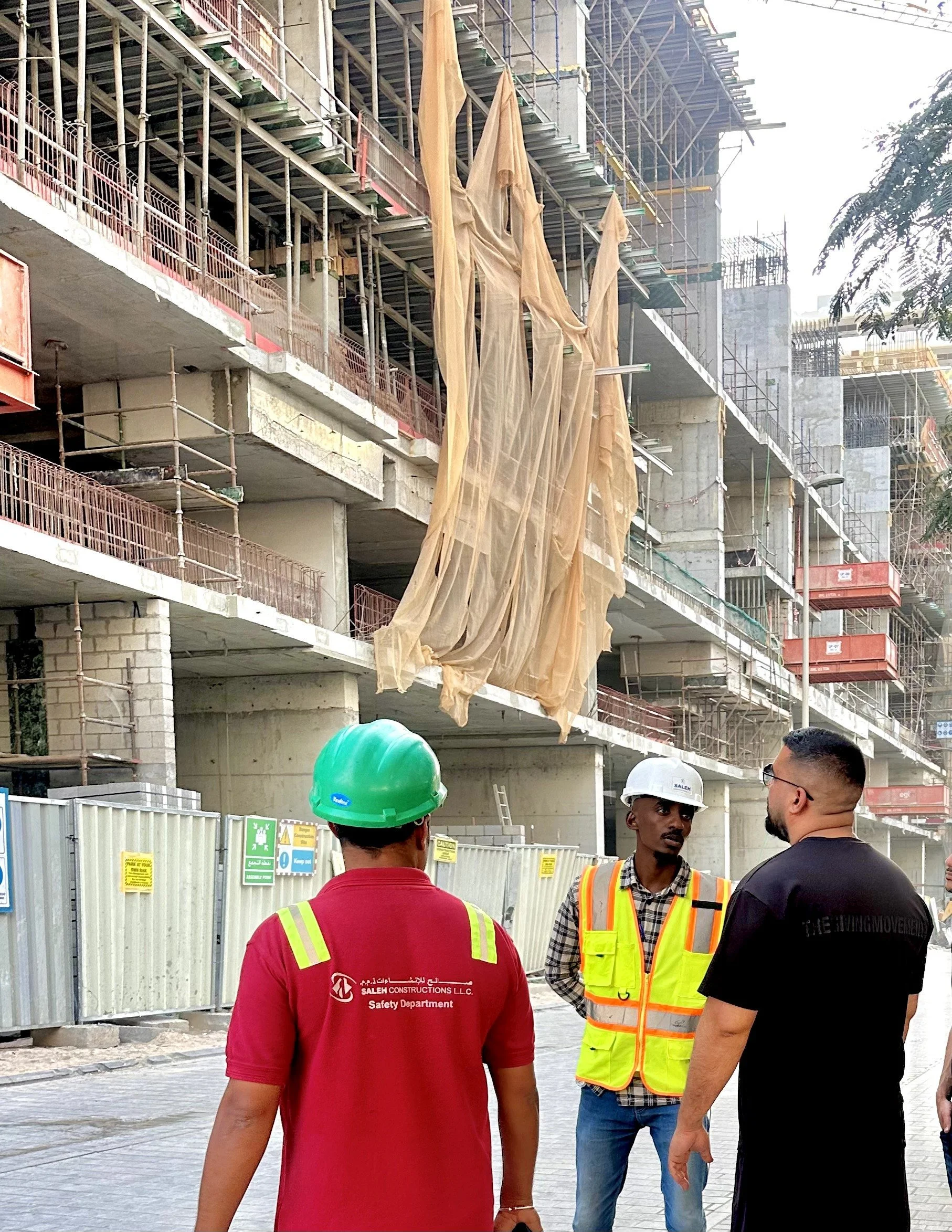 Construction workers standing in front of a building under construction, with fabric or plastic sheets hanging from the upper floors, and scaffolding around the building.