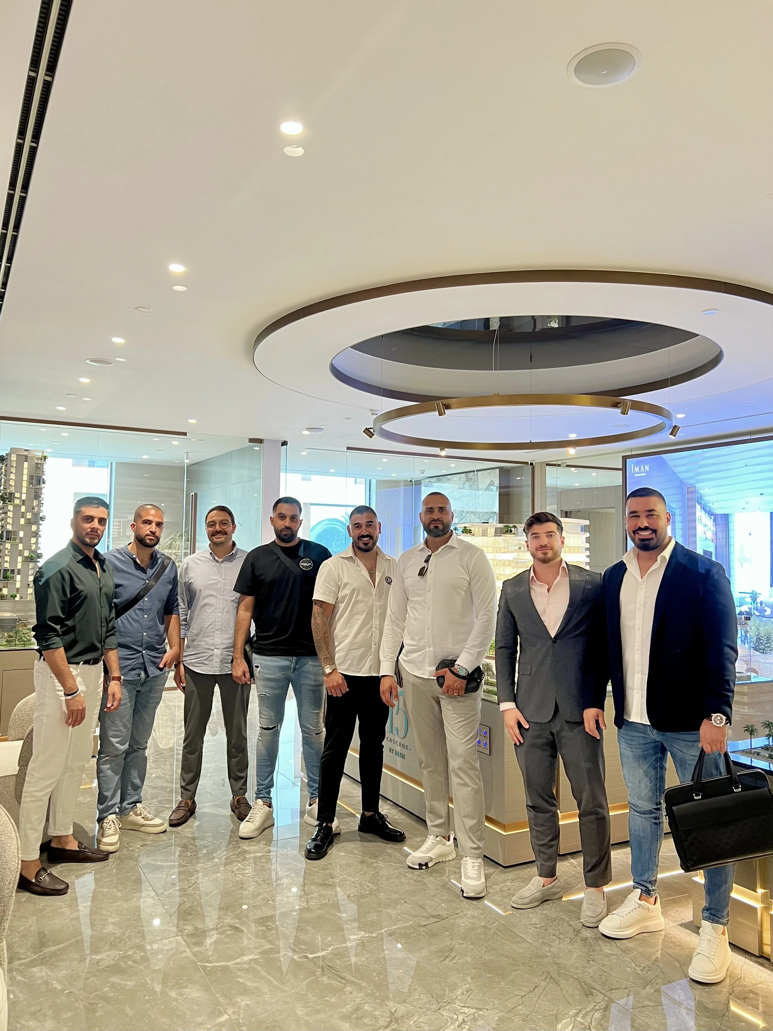 Group of nine men standing in a modern indoor setting with bright lighting, marble floor, and modern decor, some dressed in casual wear and others in suits.