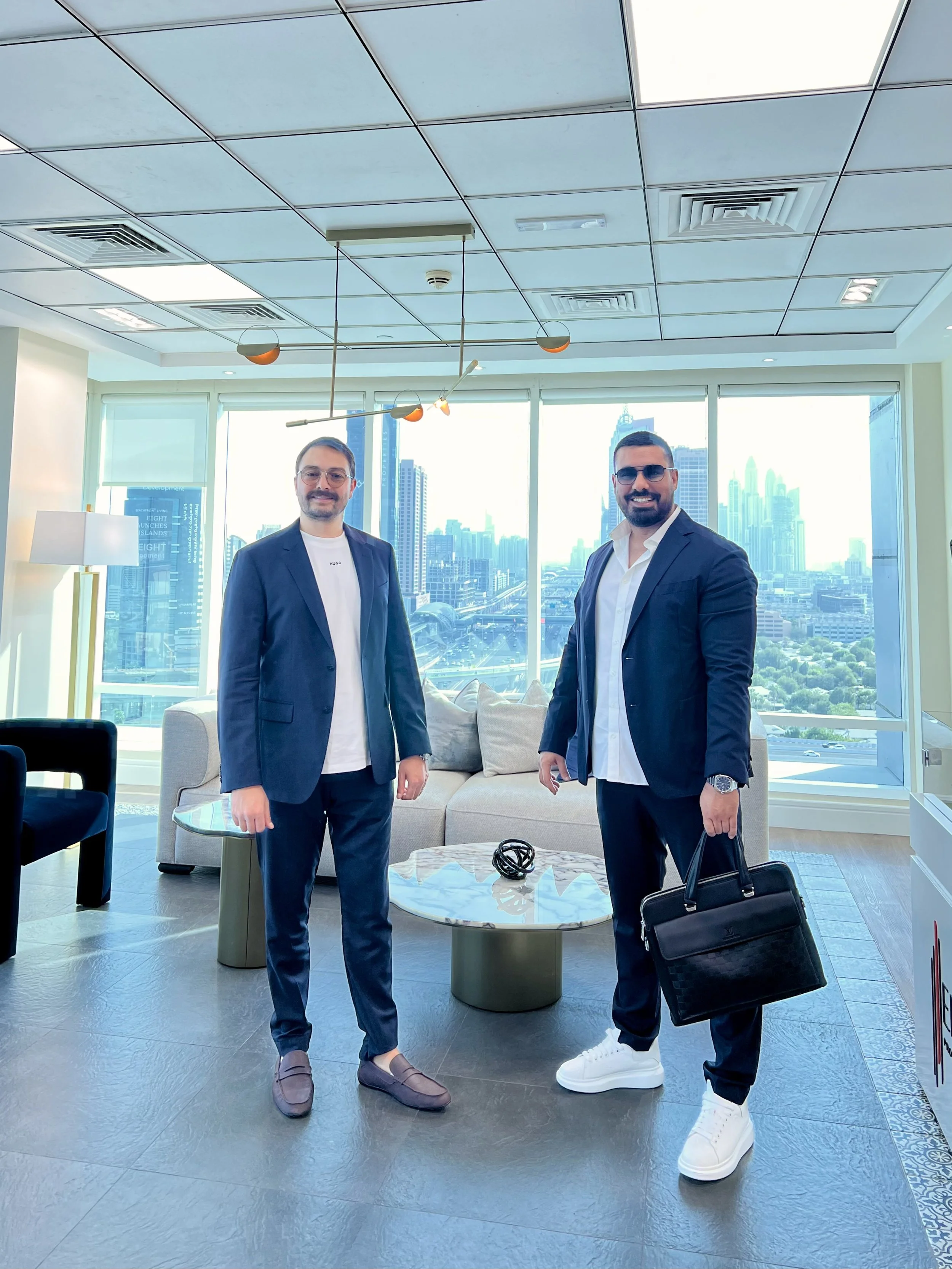 Two men in business suits standing in an office with large windows showing a city skyline, one holding a briefcase, smiling.