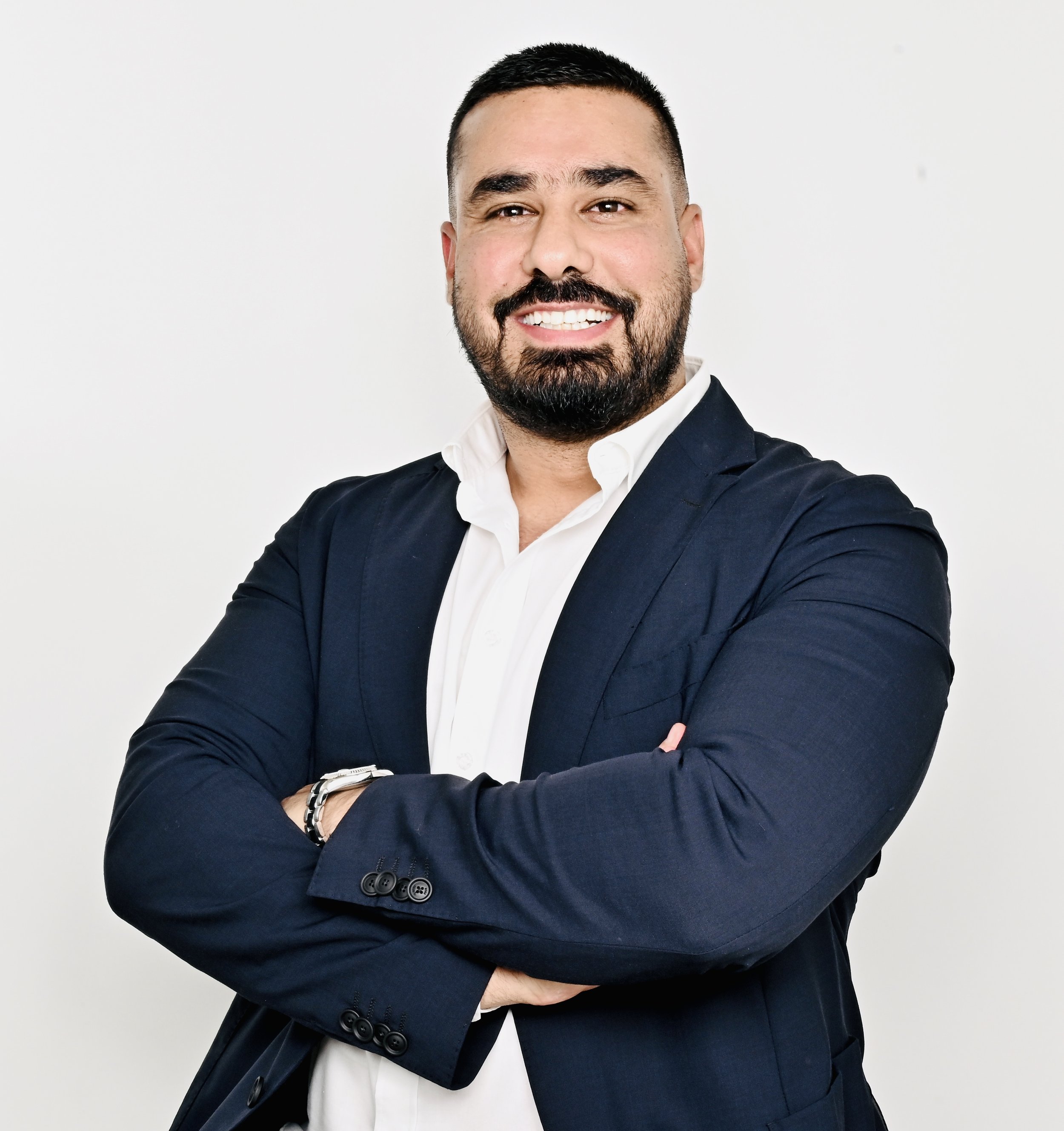 A man with a beard and short dark hair, wearing a white shirt and dark blazer, smiling with arms crossed against a plain white background.