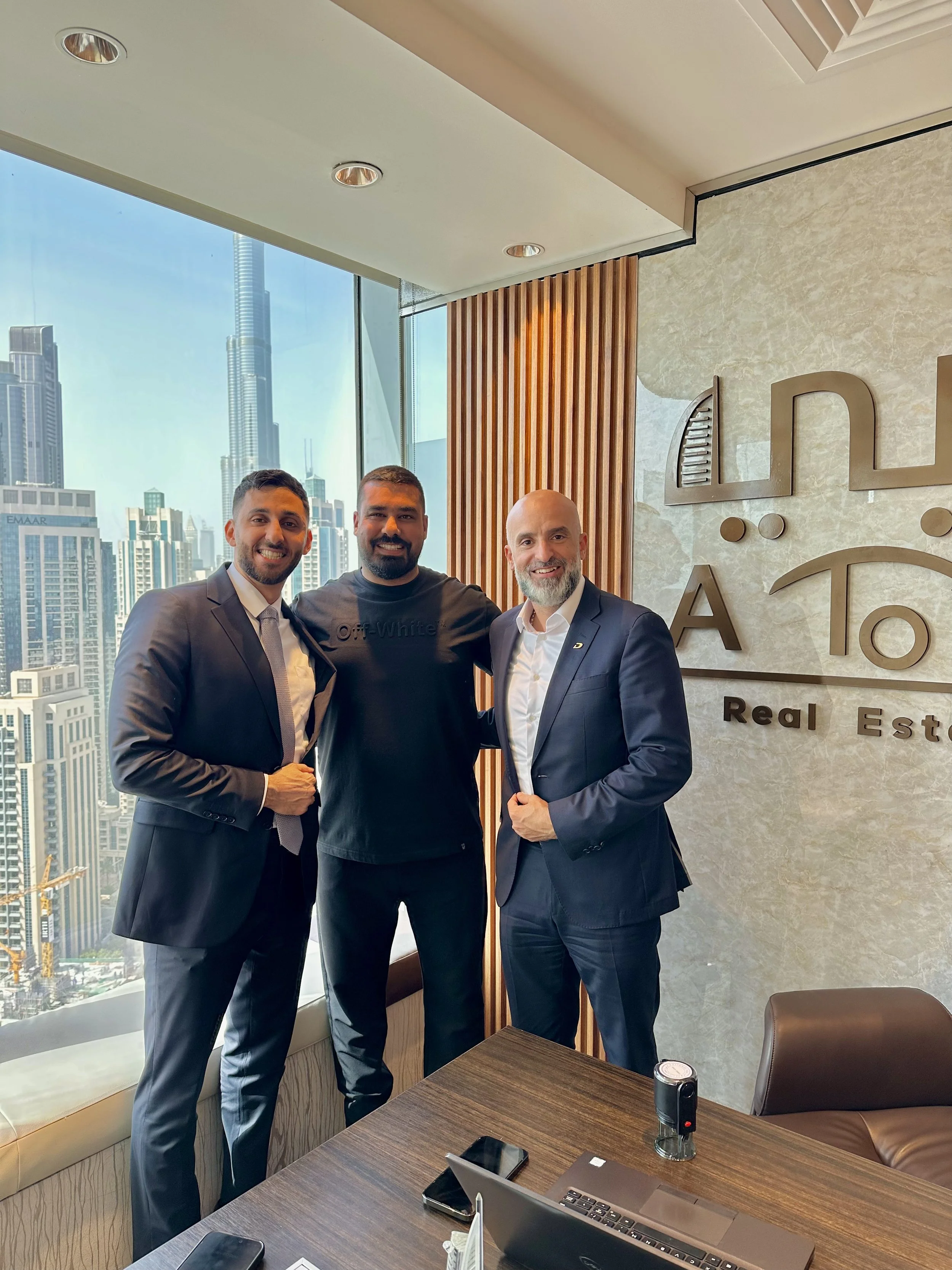 Three men in business suits and casual clothing standing inside an office with a city skyline view, including the Burj Khalifa, in the background, smiling at the camera.