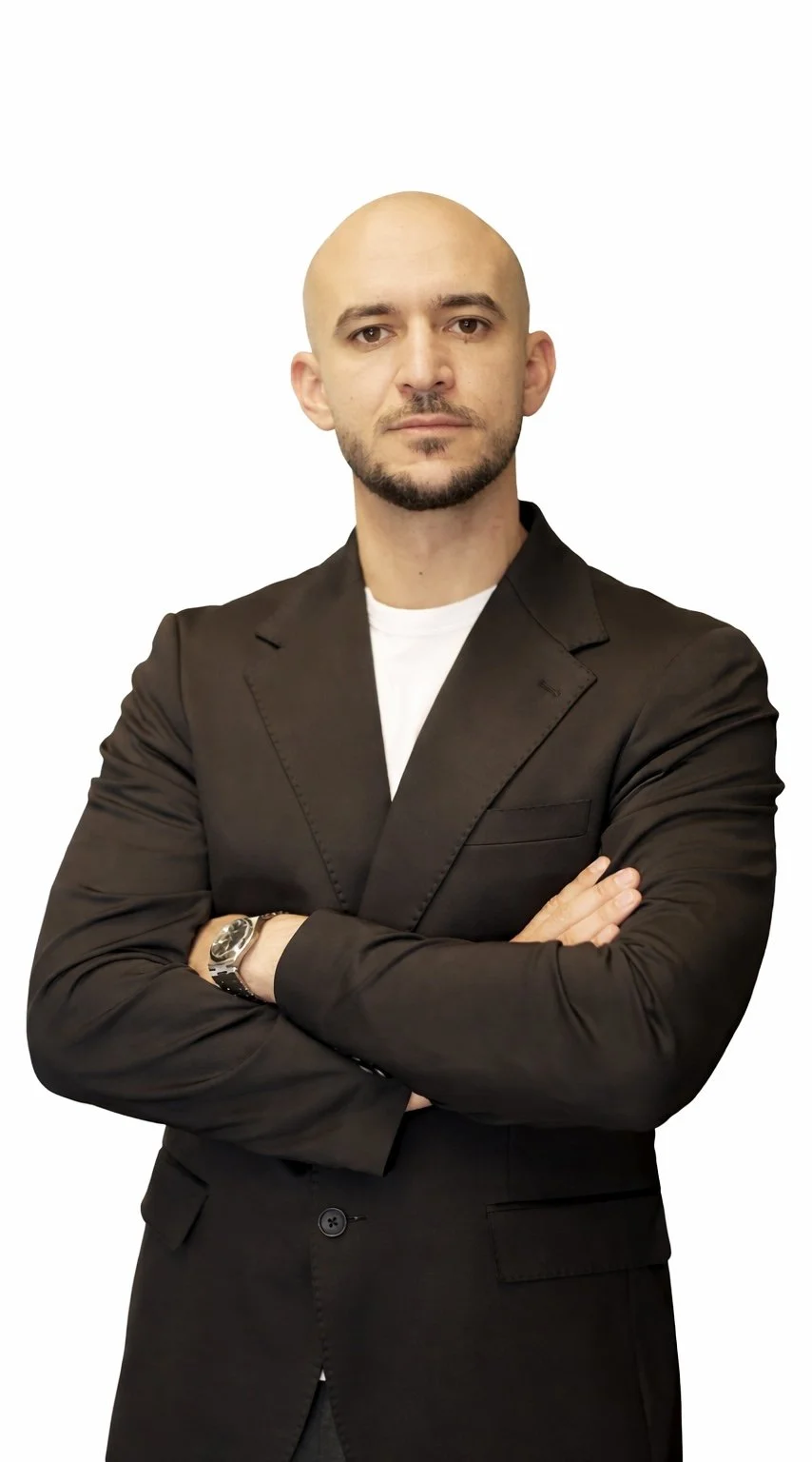 A man with a bald head and a beard, wearing a black blazer over a white shirt, standing with arms crossed against a plain white background.
