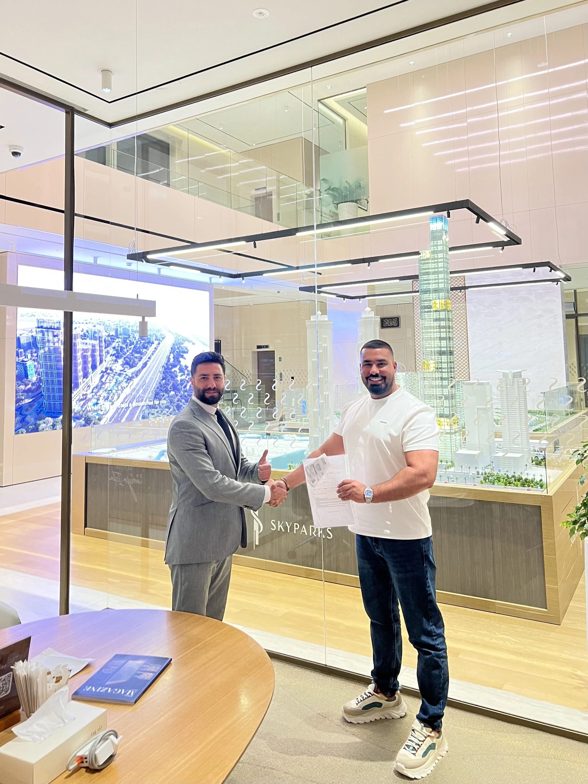 Two men shaking hands inside a building, with one in a suit and the other in casual clothes, in front of a model of skyscrapers labeled 'Skyparks'.