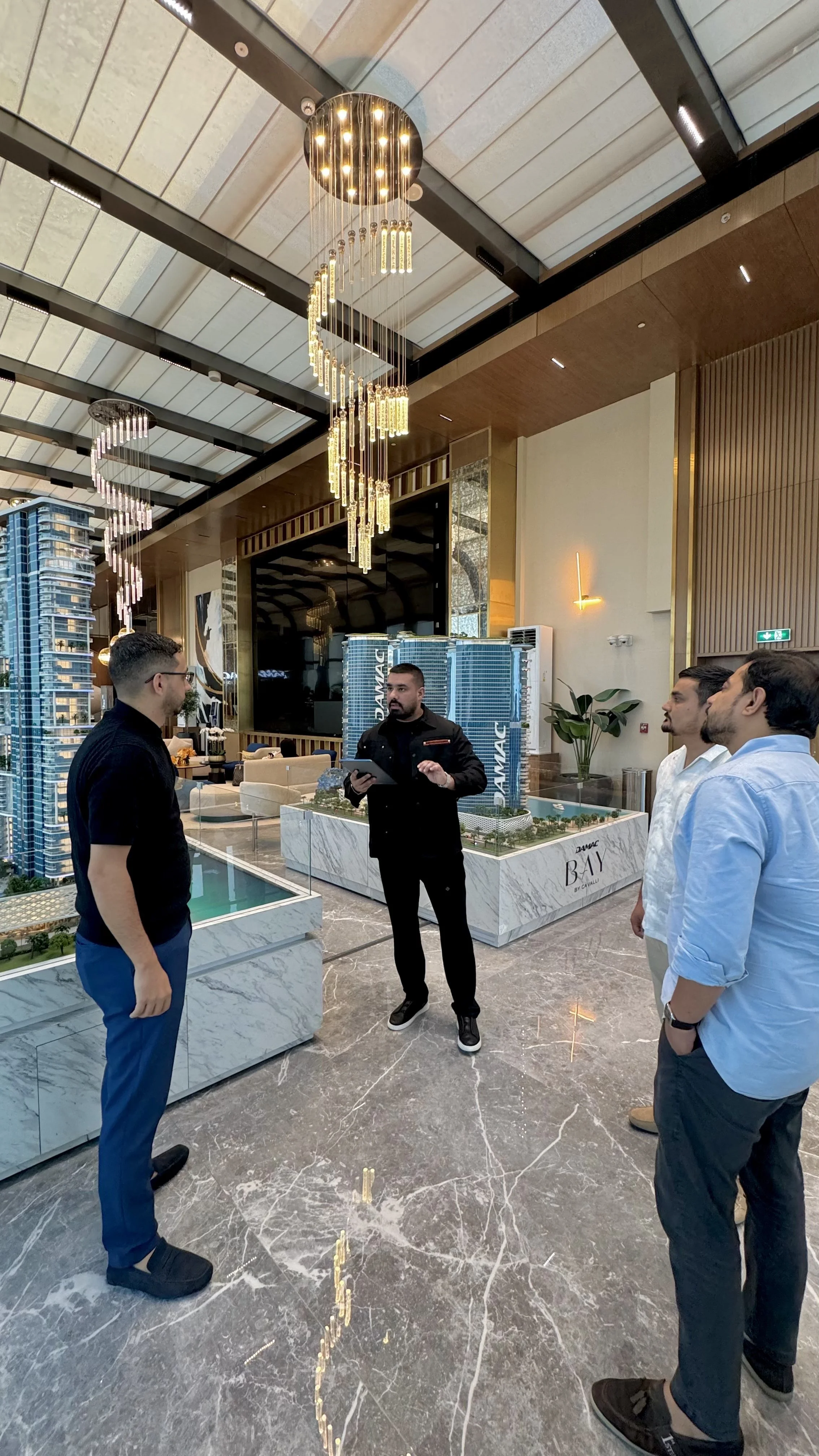 Four men are standing in a luxury real estate sales office, engaging in conversation while looking at a scale model of a real estate development. The office features modern design elements, high ceilings with decorative chandeliers, and large windows