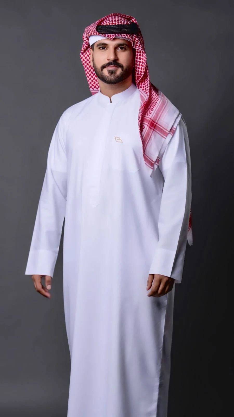 Man wearing a traditional white thobe and red and white checkered keffiyeh against a gray background.