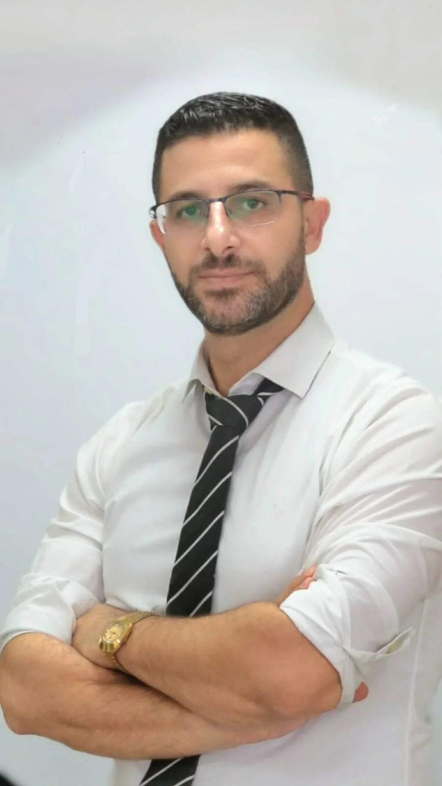 A man with glasses, dark hair, and a beard, wearing a white dress shirt, a striped black tie, and a gold wristwatch, standing with arms crossed in front of a plain white wall.