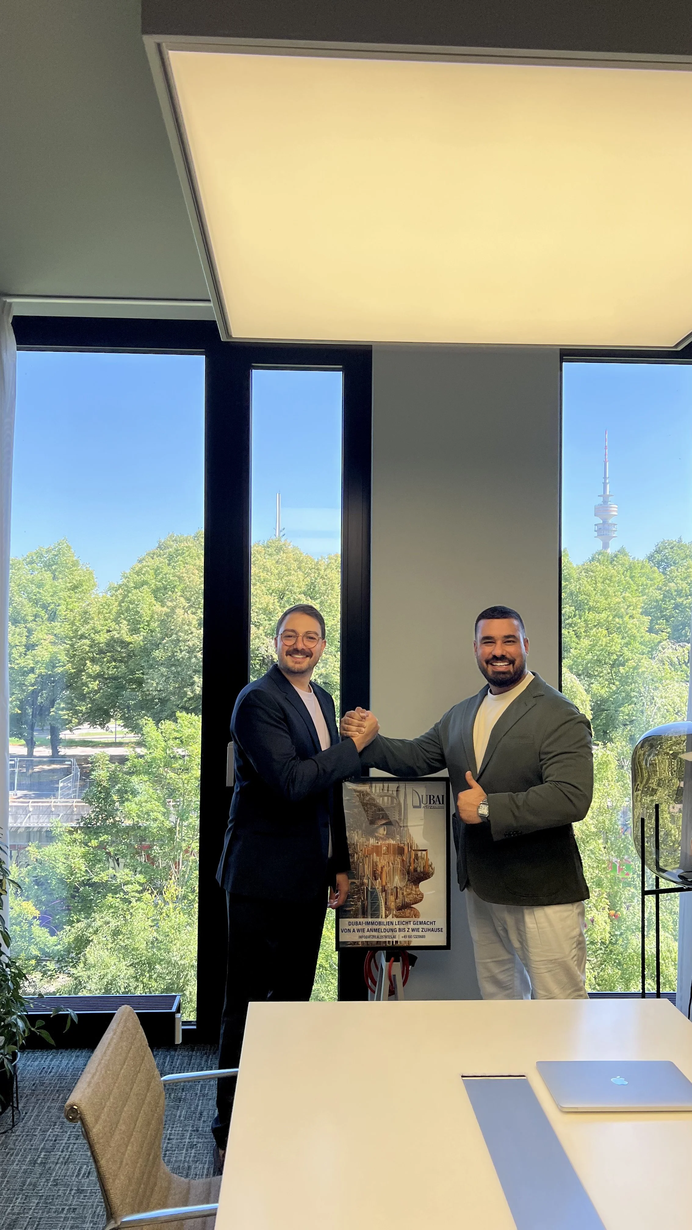 Two men in business attire shaking hands and smiling inside an office, with large windows showing green trees and a cityscape with a tall tower in the background.