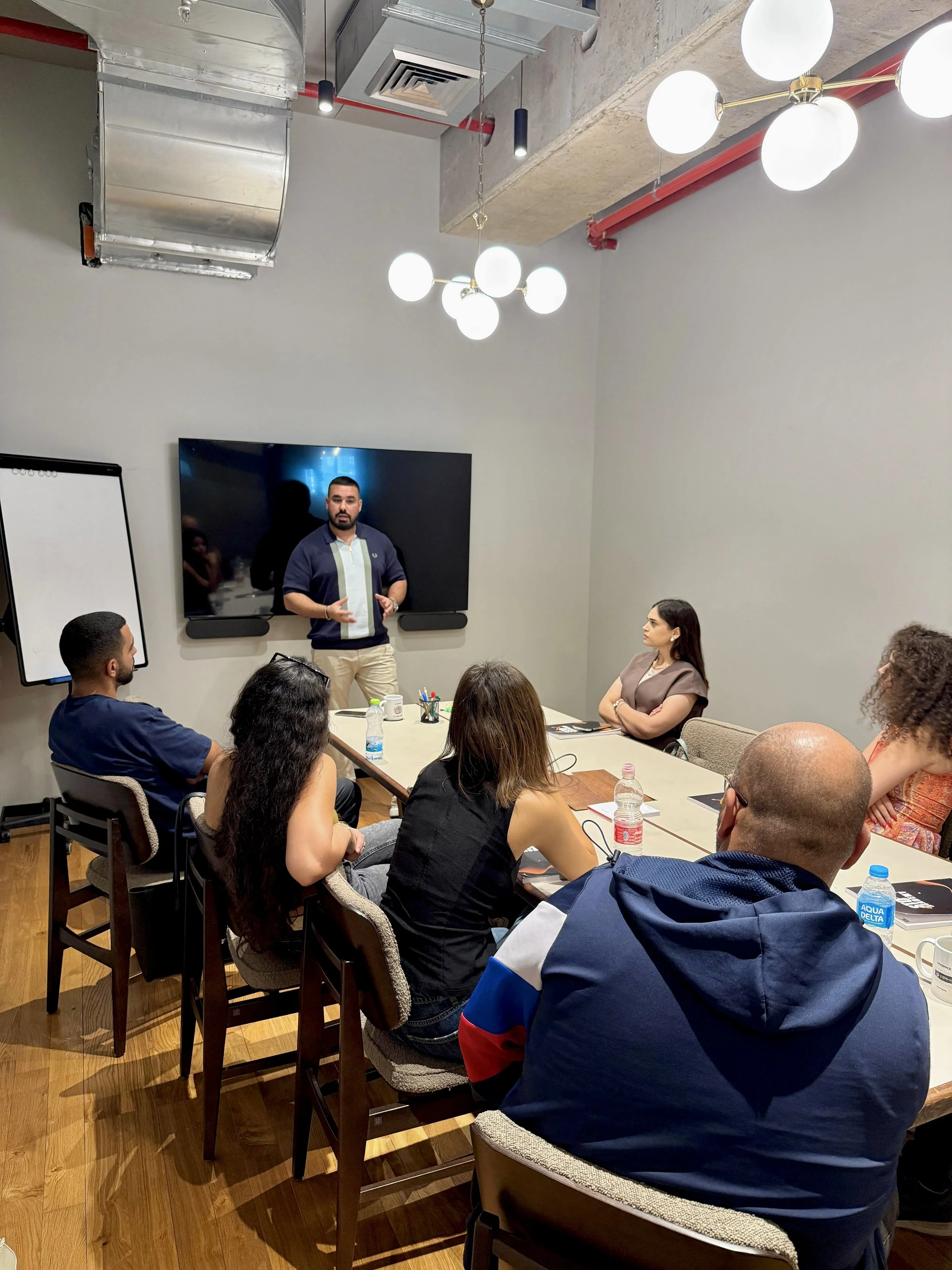 A man giving a presentation to a group of six people in a conference room with wooden flooring, white walls, and modern lighting fixtures.