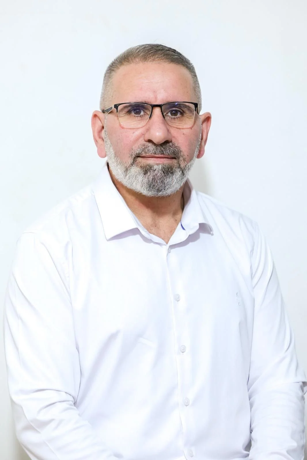 A middle-aged man with glasses, a beard, and short hair, wearing a white collared shirt, standing against a plain white background.