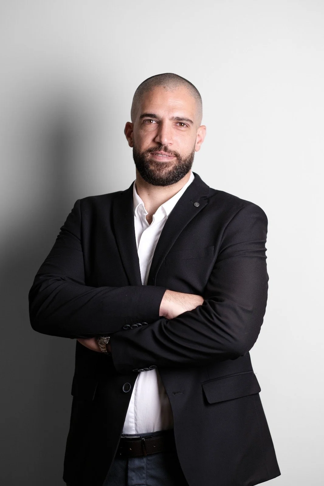 A man with a shaved head and beard wearing a black blazer over a light patterned shirt standing against a plain, light-colored wall.