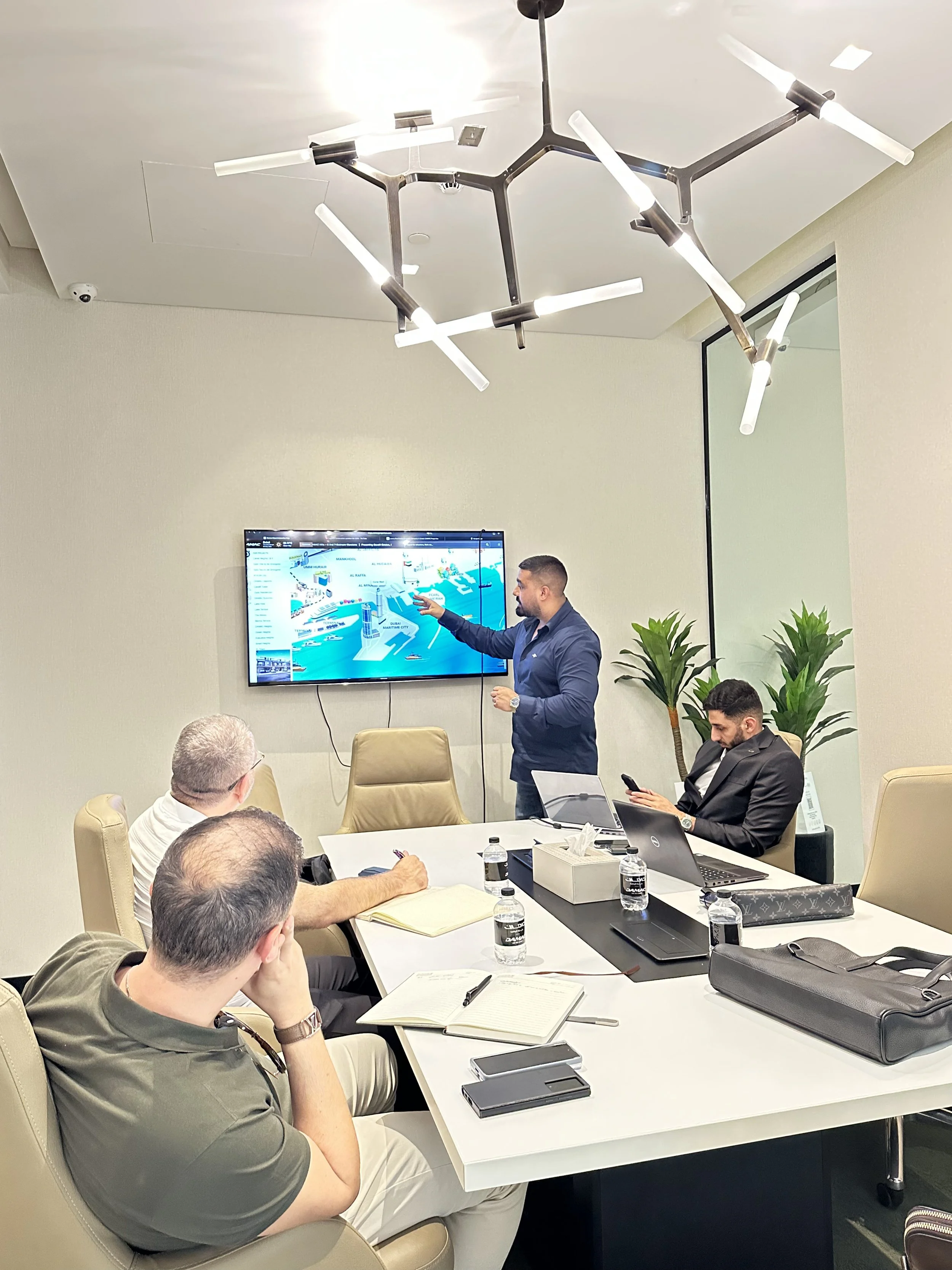 Business meeting in a conference room with four men, one standing and presenting on a screen, others seated taking notes or working on laptops, four water bottles on the table, modern light fixture on ceiling, potted plants in the corner.