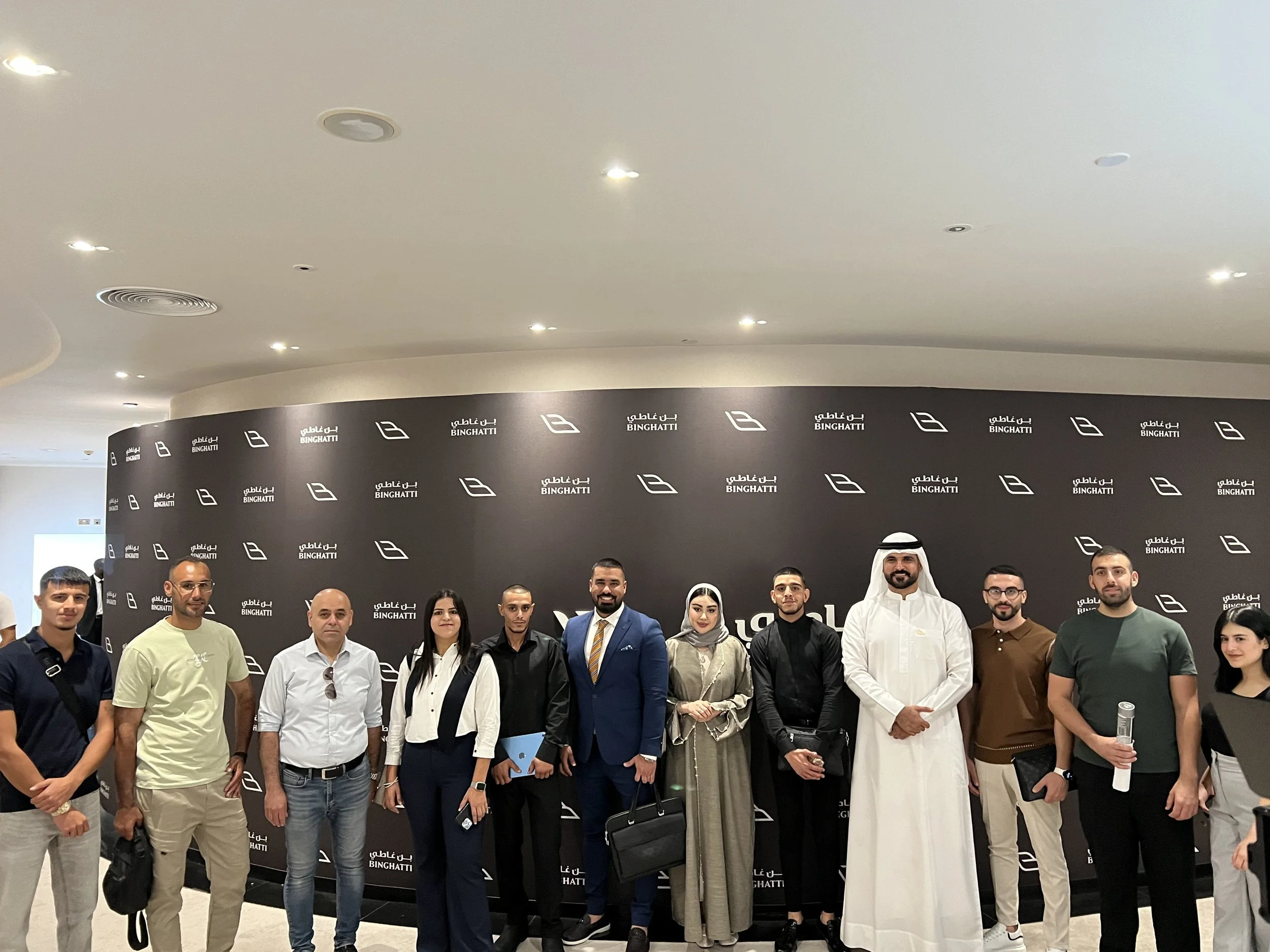 Group of fifteen people posing in front of a black backdrop with white logos and text, in an indoor setting with ceiling lights.