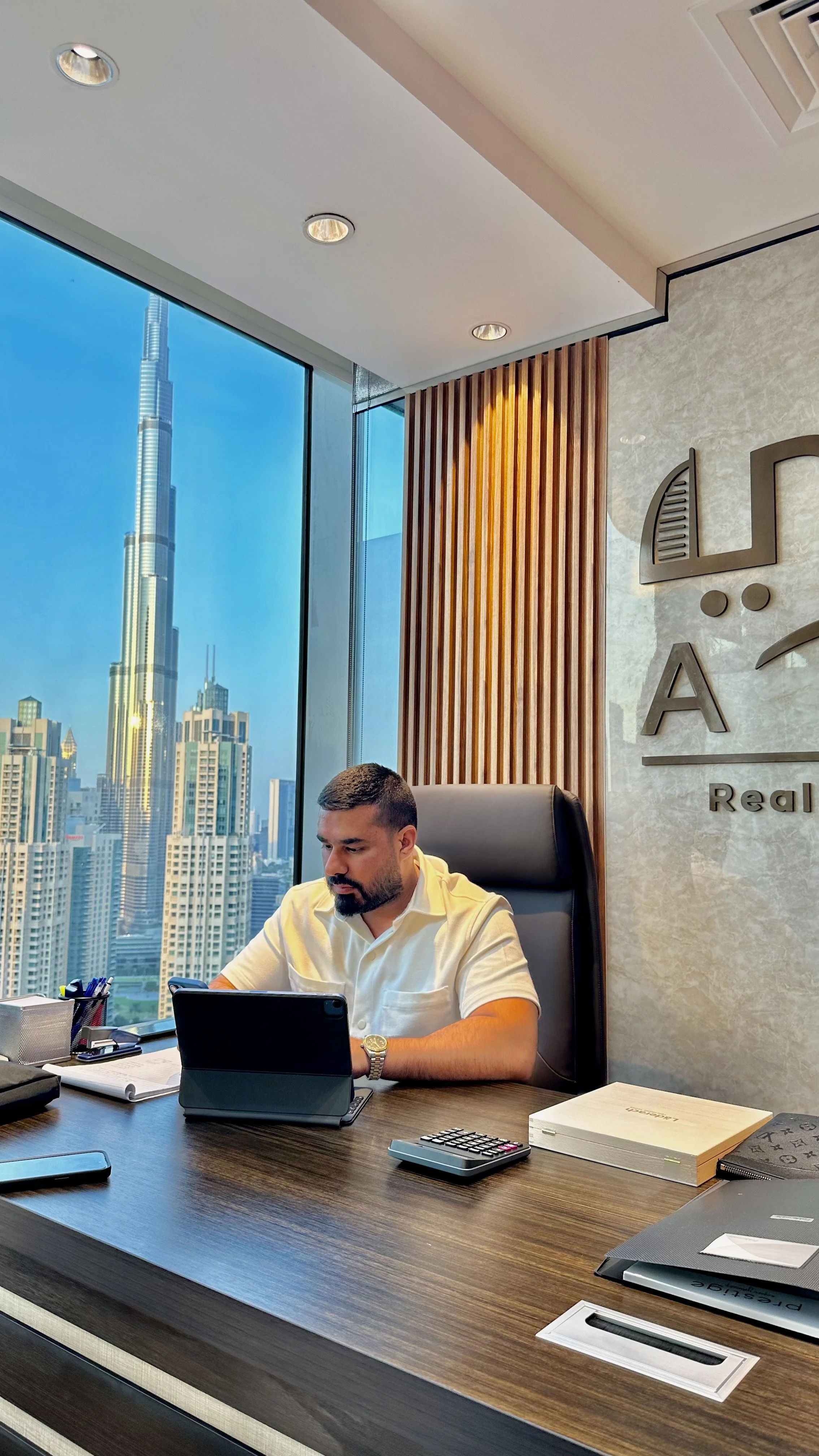 A man sitting at a desk in a modern office with a view of Dubai's skyline, including the Burj Khalifa, working on a tablet.