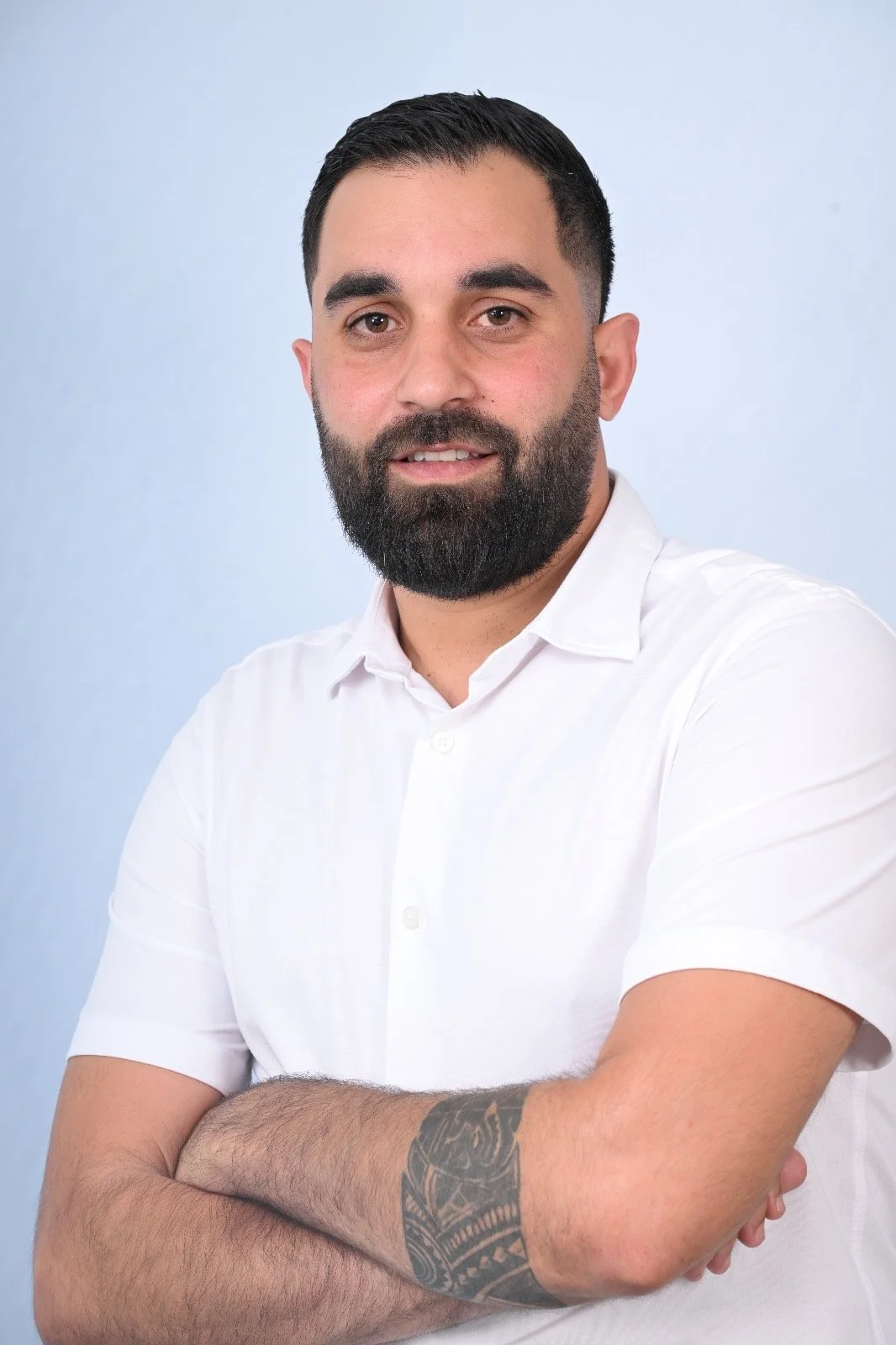 Portrait of a man with a beard, wearing a white shirt, crossing his arms, against a light blue background.