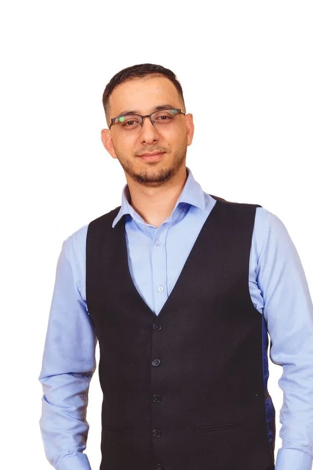 Man in glasses wearing a blue shirt and black vest, standing against a white background.