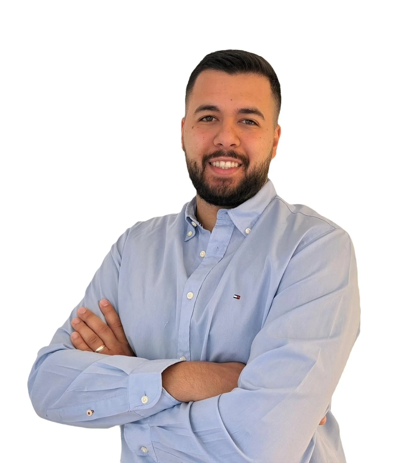 A man with short dark hair and a beard, smiling and crossing his arms, wearing a light blue button-up shirt, standing against a plain off-white wall.