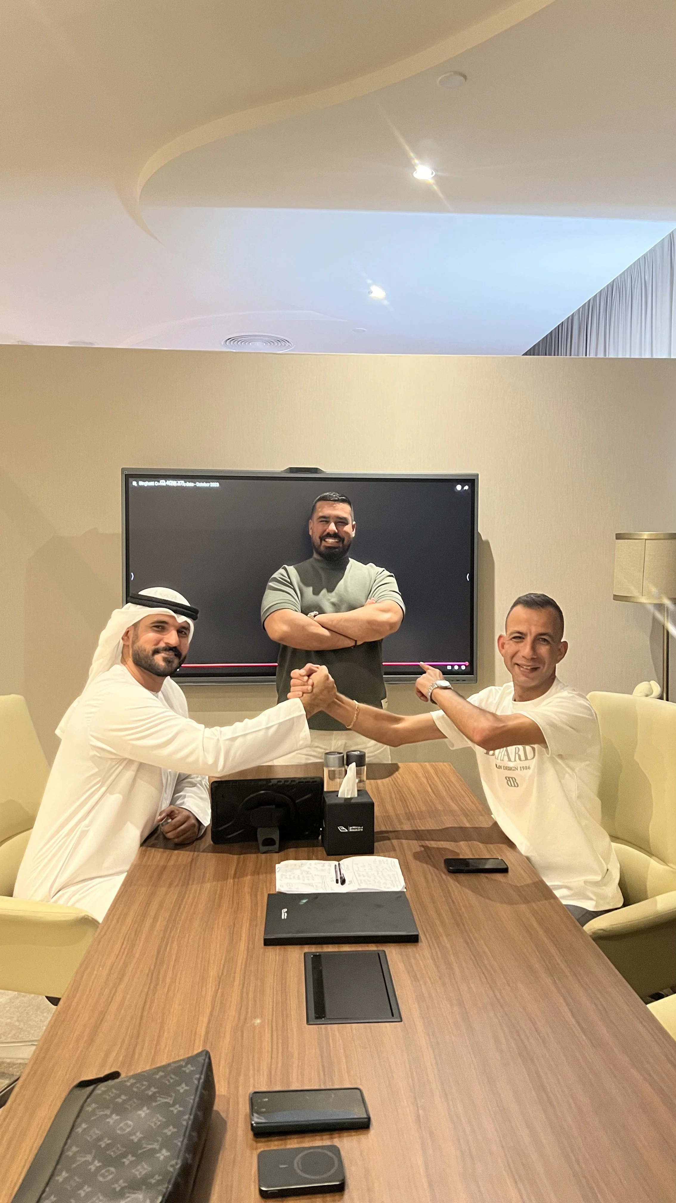 Three men in a conference room, two sitting at a wooden table and shaking hands, and one standing in the background with arms crossed, smiling. The table has notebooks, a phone, and a conference phone. Behind the standing man is a large screen.