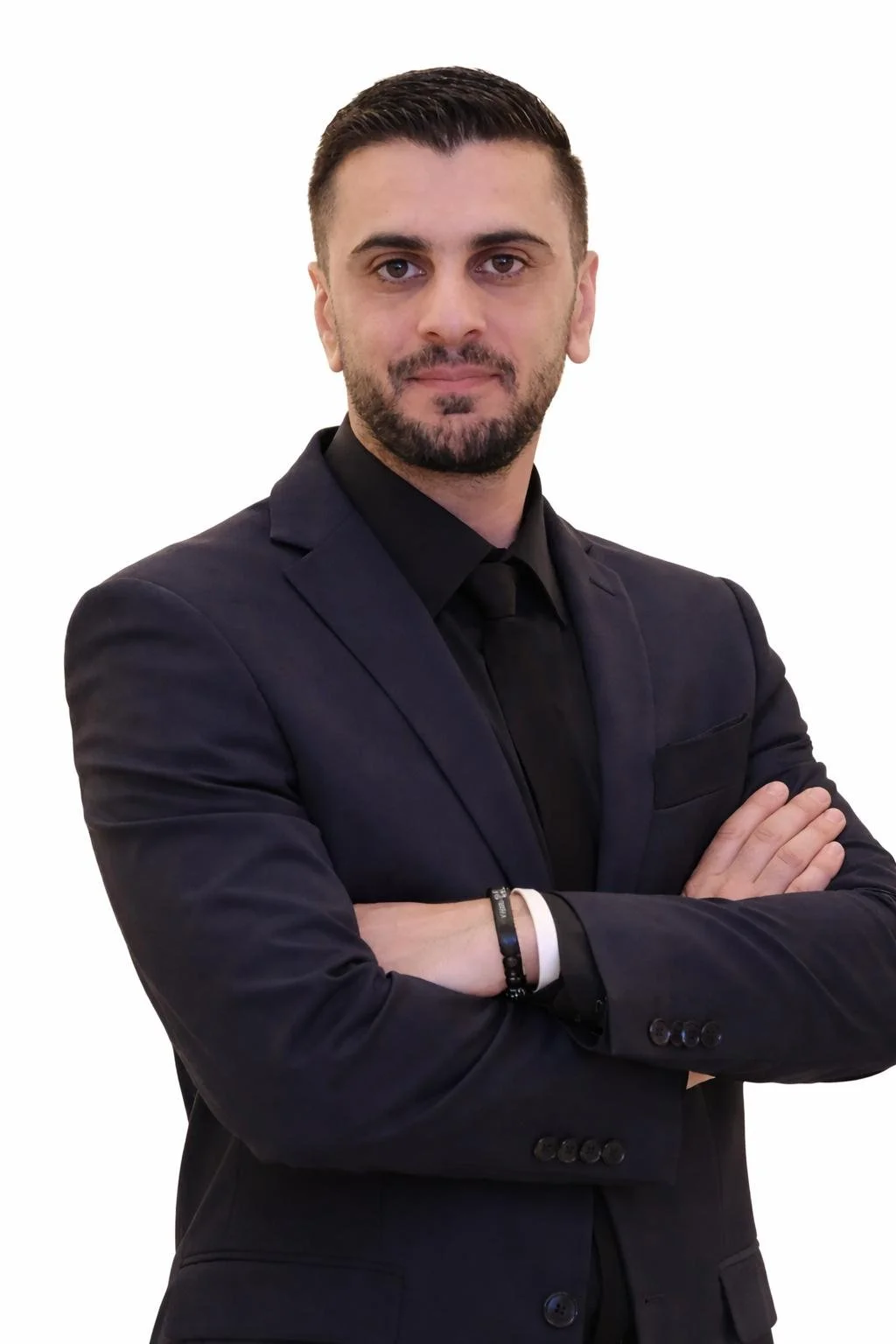 A man with dark hair and a beard, wearing a black suit, black shirt, and black tie, standing with arms crossed against a plain white background.