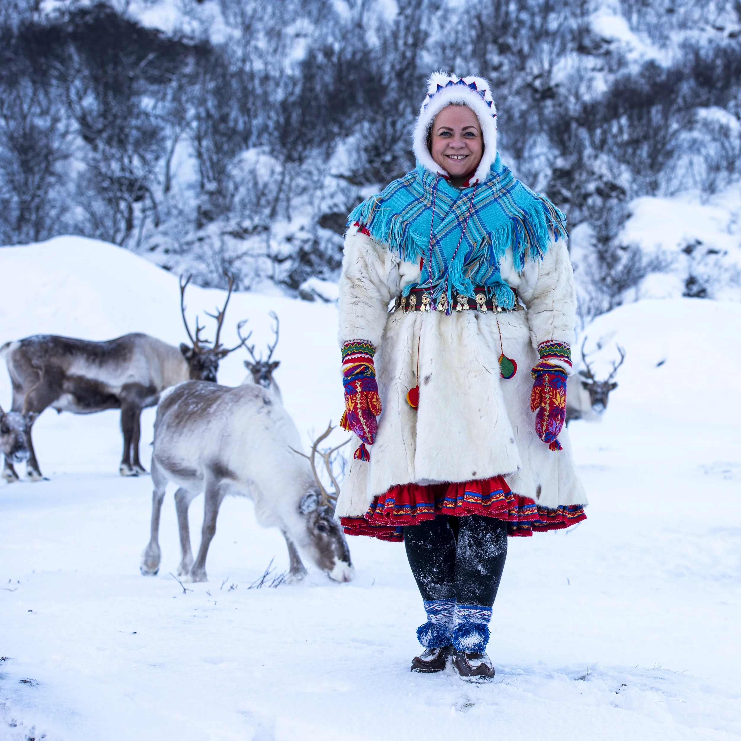 En person i tradisjonell samisk drakt som står i snøen med reinsdyr i bakgrunnen.