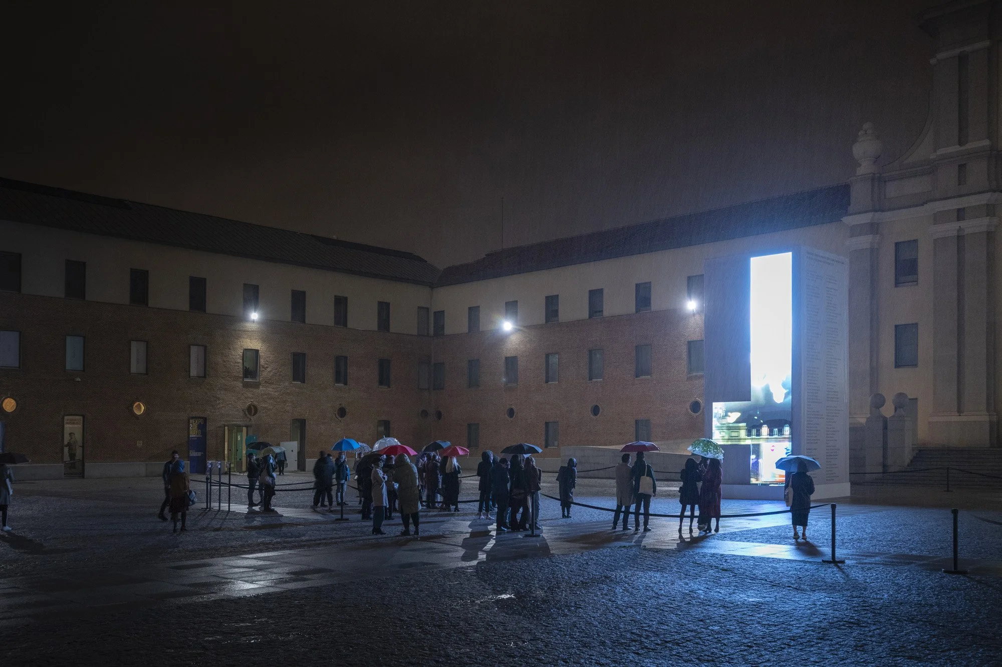 Personas con paraguas en una fila frente a una pantalla grande en una plaza en noche lluviosa.