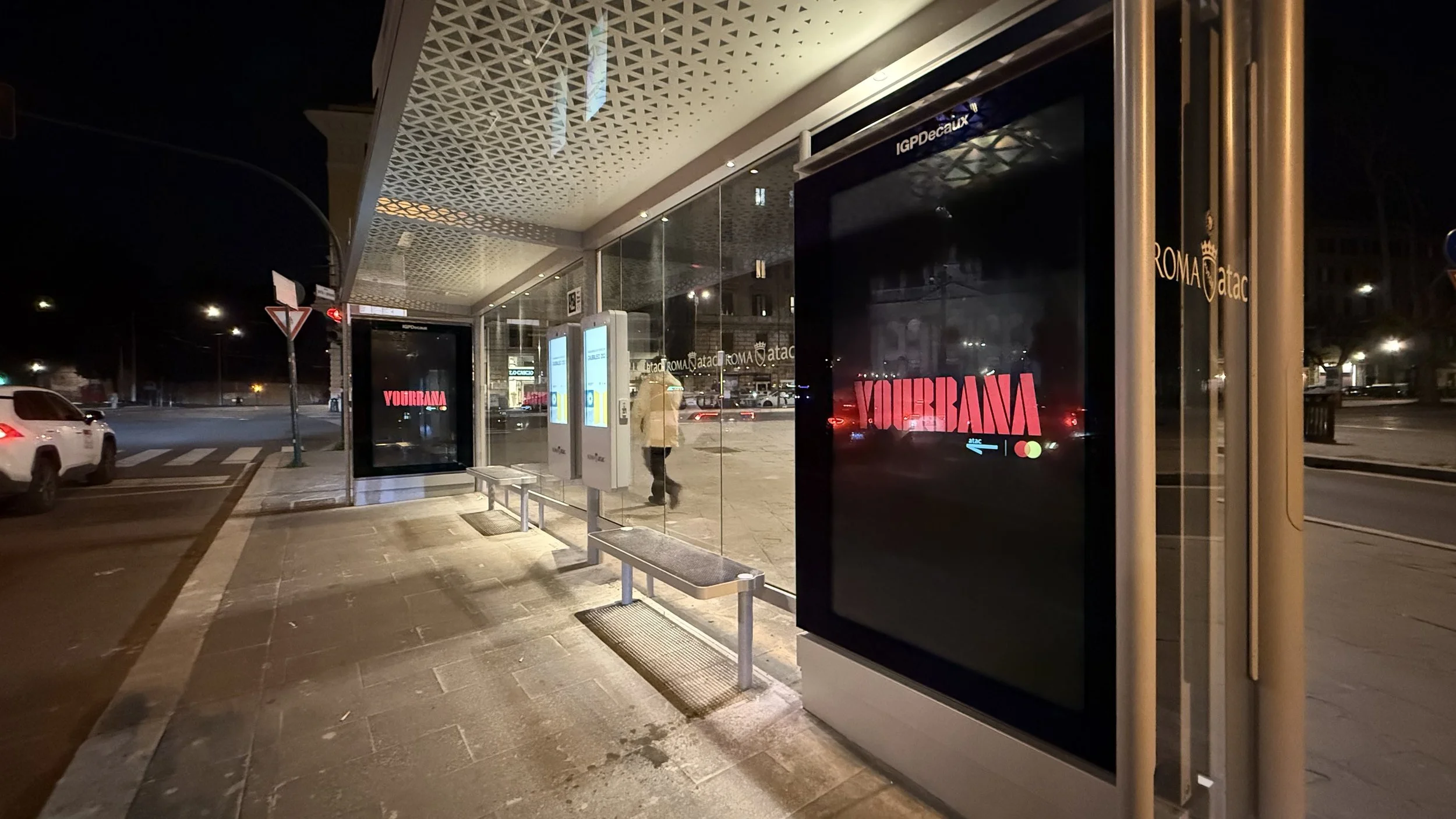 Parada de autobús en la noche con carteles y una pantalla digital que muestra la palabra 'Yucatán' en letras rojas, en una calle con vehículos y una persona caminando.