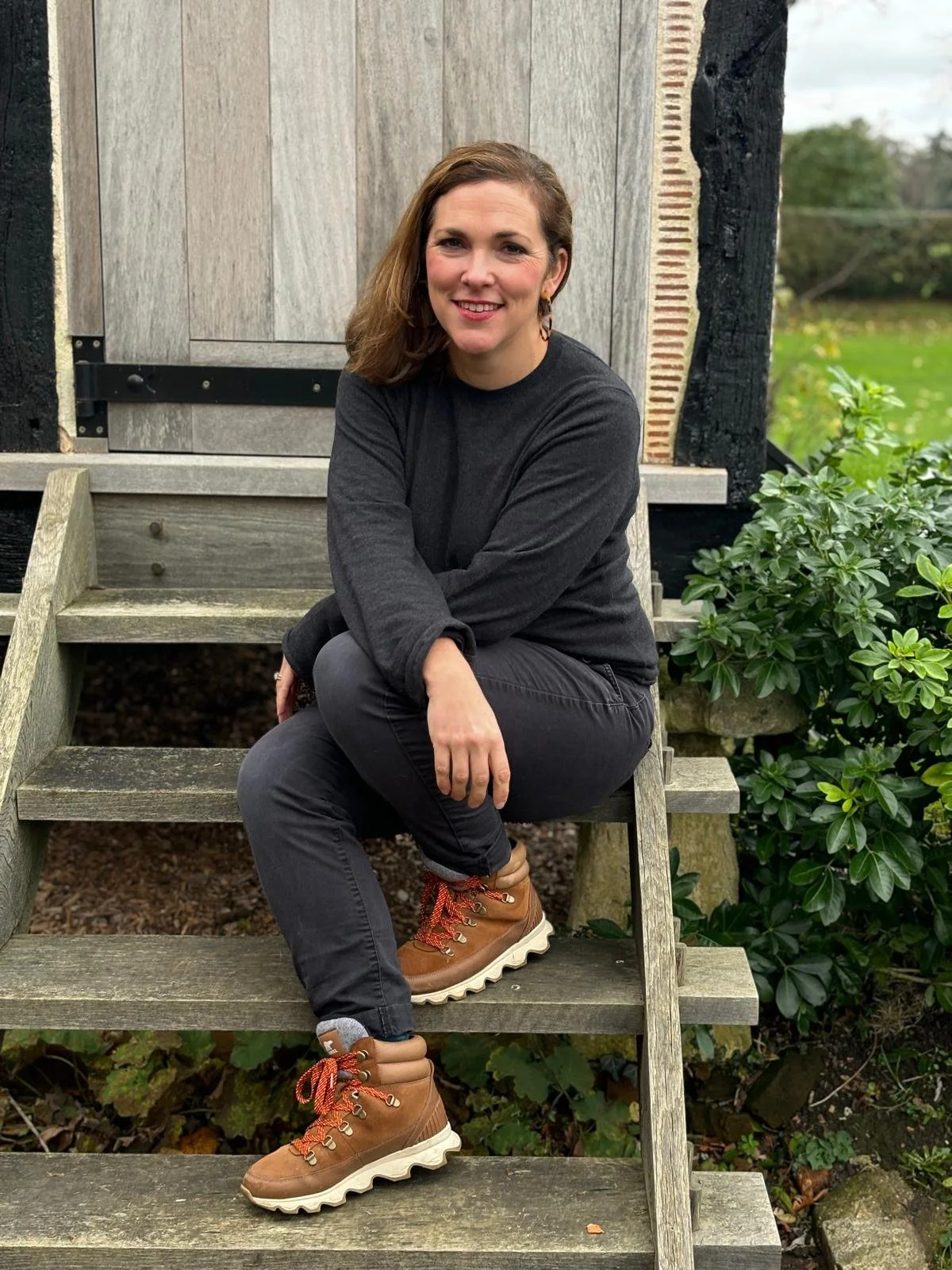 A woman with brown hair sitting on wooden stairs outdoors, wearing a dark grey long-sleeve shirt, black pants, and tan hiking boots, smiling at the camera with a garden and shed in the background.