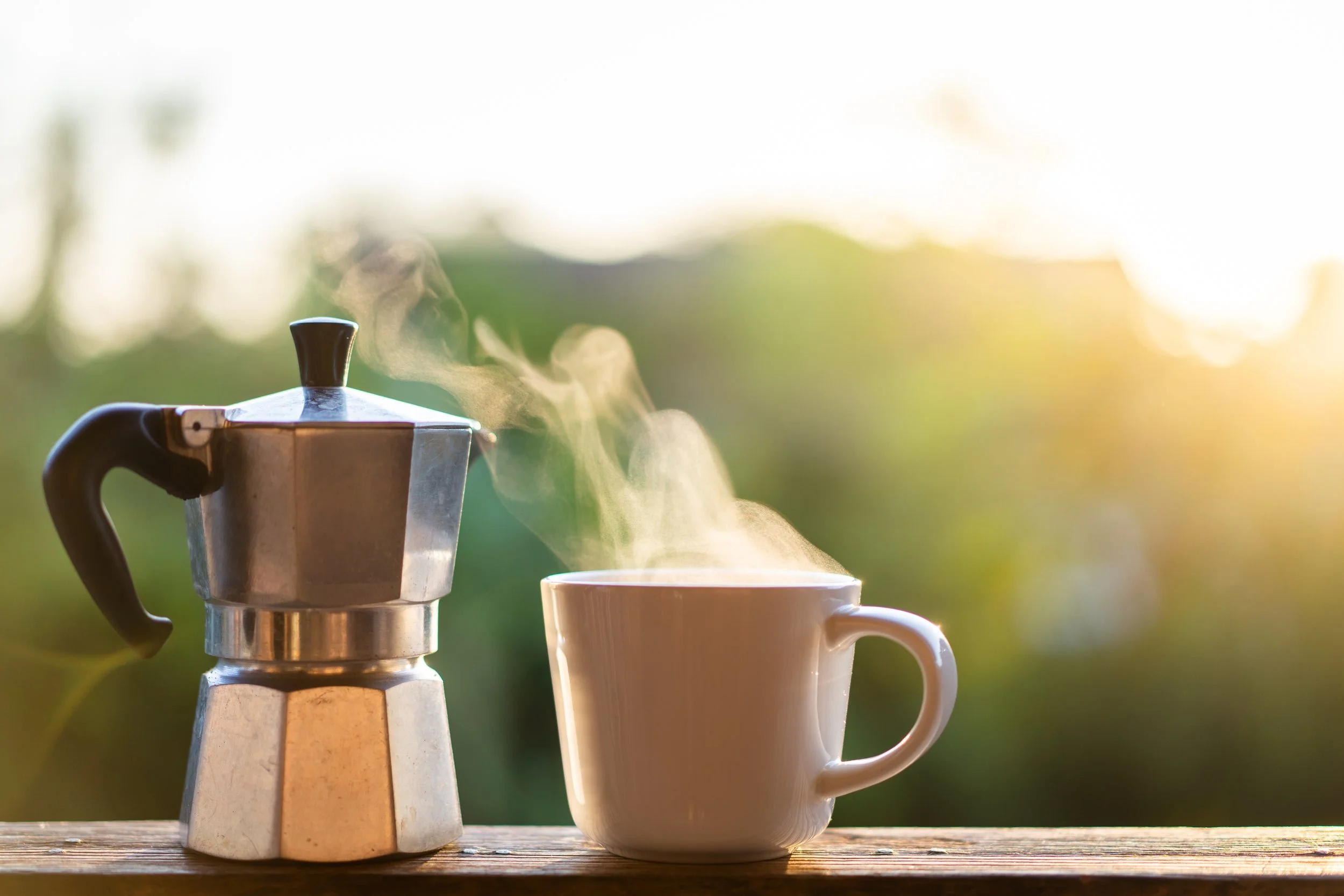 A stovetop espresso maker next to a steaming white mug on a wooden surface outdoors during sunrise or sunset.