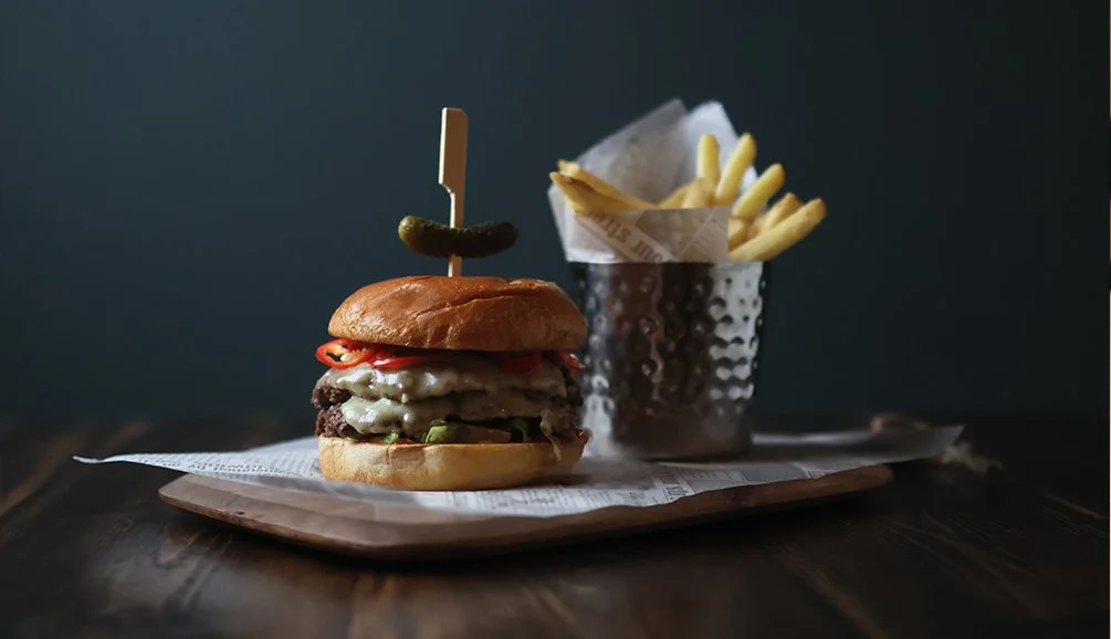 A cheeseburger with pickles, tomato, lettuce, cheese, and a beef patty in a bun with a pickle spear inserted, served with a side of French fries in a metal cup.
