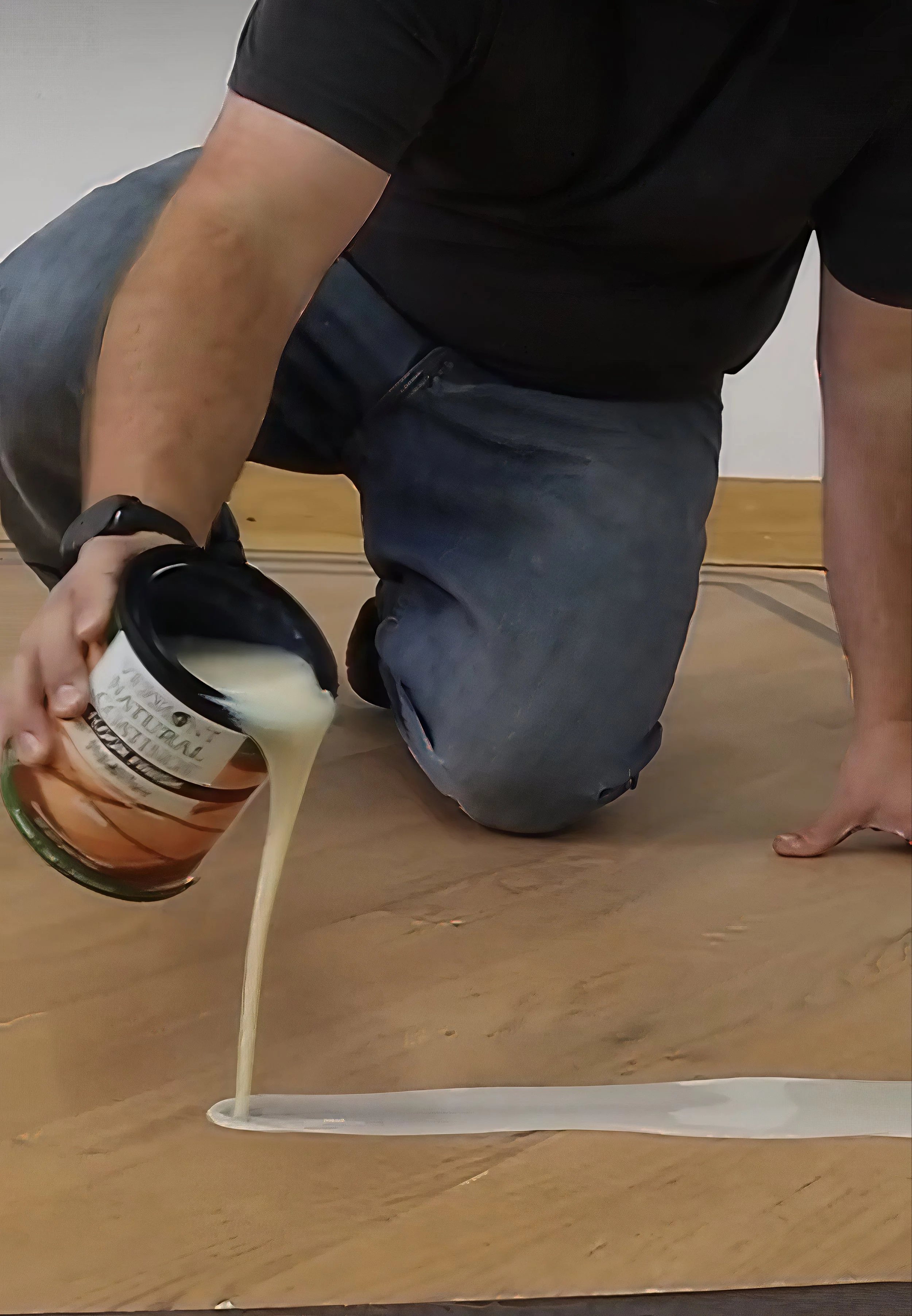 A person kneeling on a wooden floor is pouring a thick, white substance from a jar onto the floor, creating a long streak.