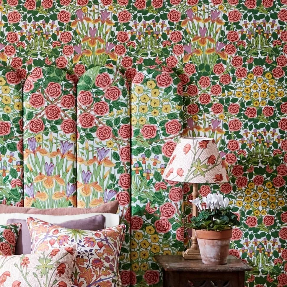 A bedroom with vibrant floral wallpaper featuring pink roses, yellow flowers, green foliage, and purple irises. There are decorative pillows with floral patterns on the bed. A wooden nightstand holds a potted white flower plant and a table lamp with a floral lampshade.